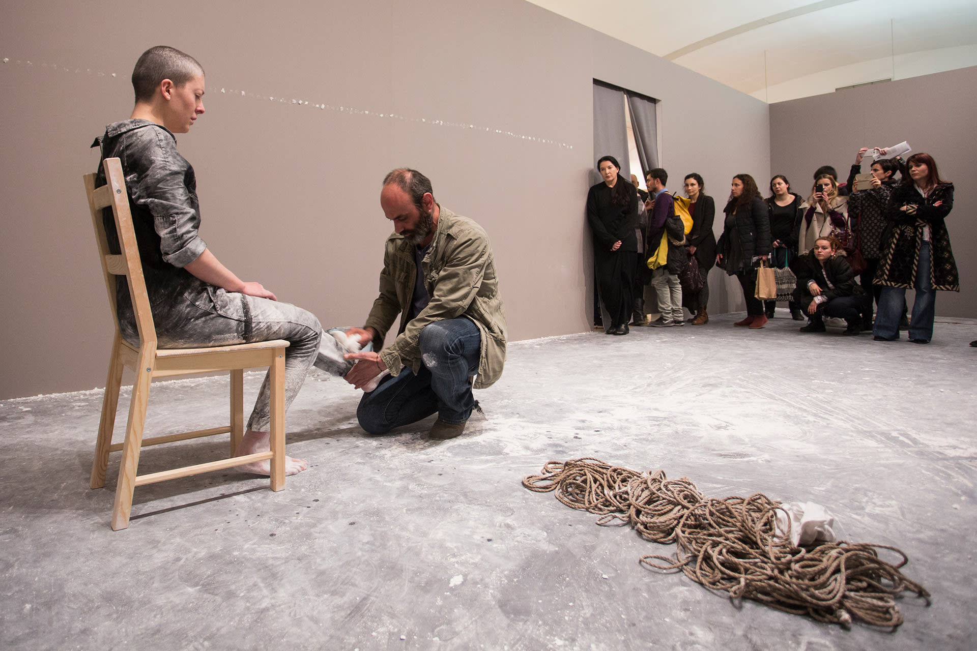 Shaved-head female performer in a worker's uniform, in bondage, sitting on a chair, having her feet wiped by a visitor.