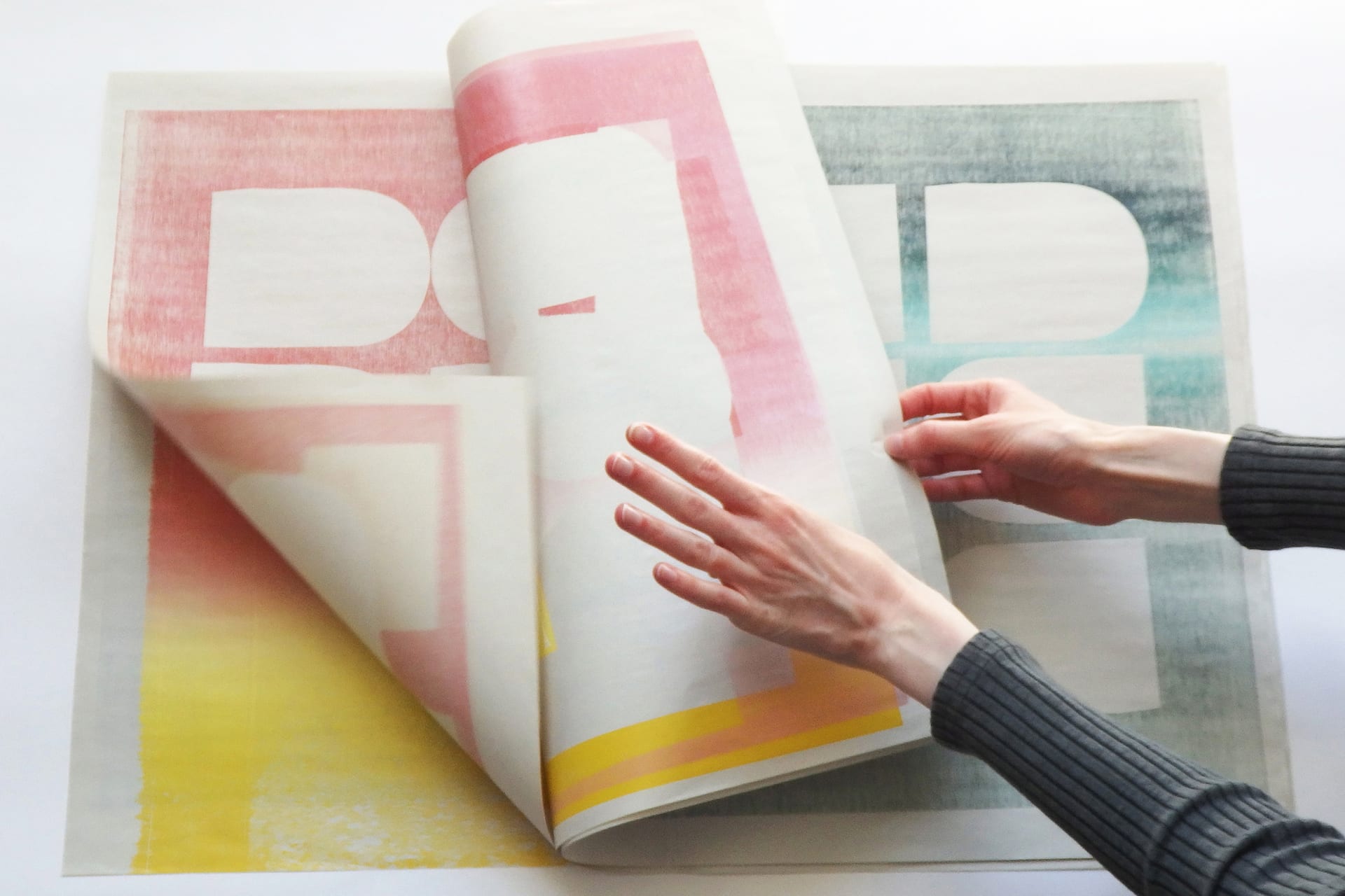 Pages of a book being flipped through, showing woodblock prints of cut out shapes and yellow, pink and green gradients.