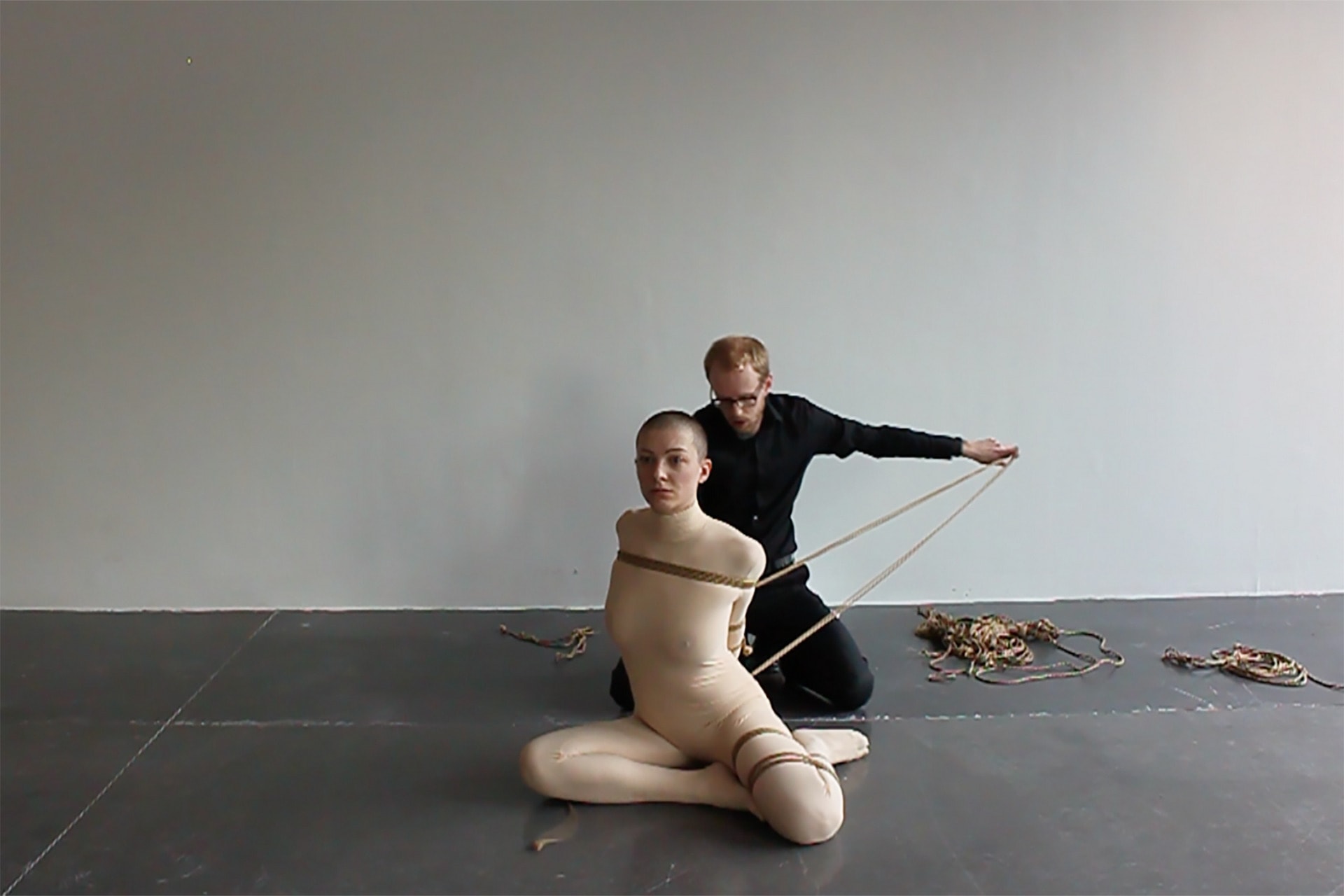 Shave-head female performer, wearing a skin-toned full body-suit, in bondage being untied by a male rigger in black. 