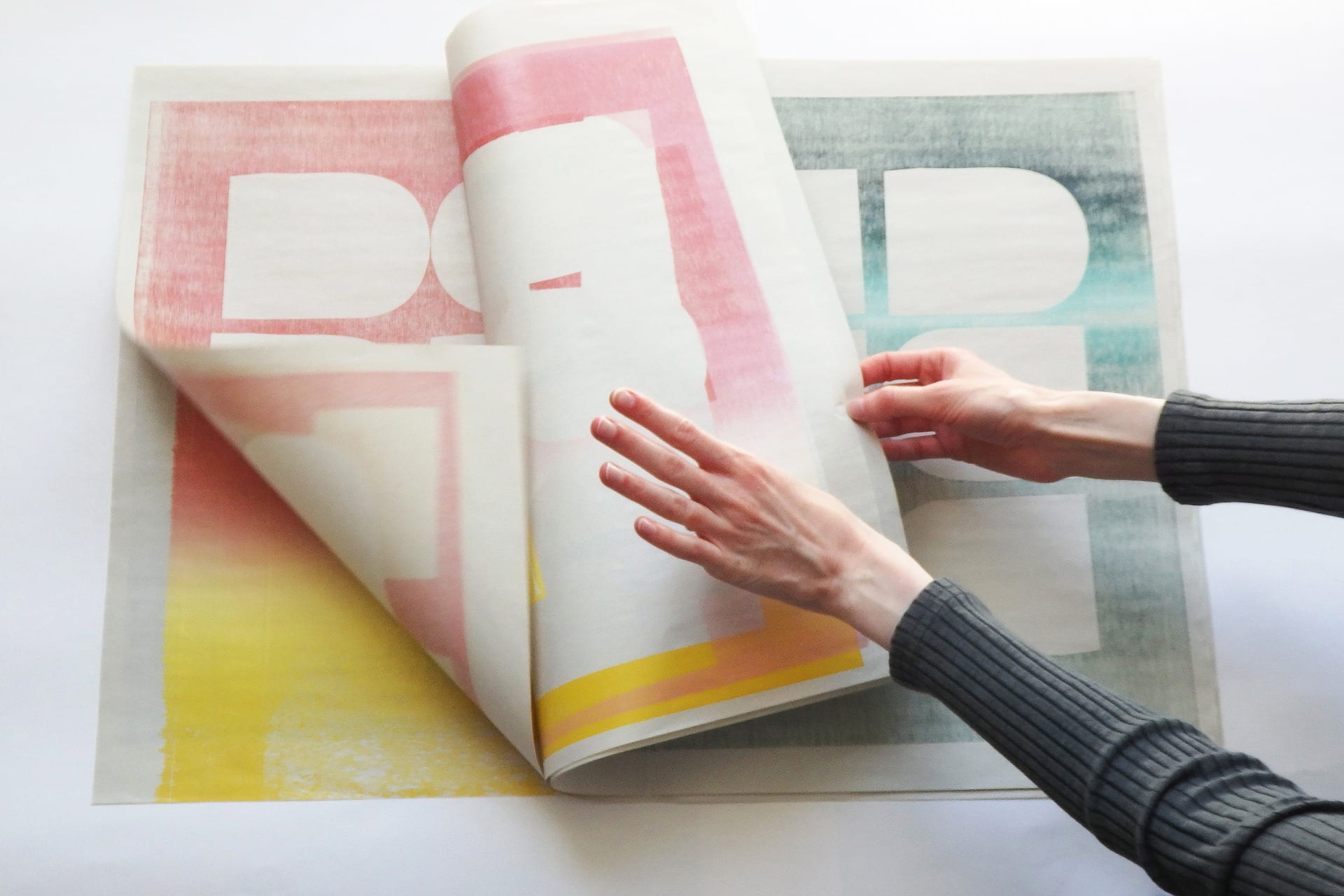 Pages of a book being flipped through, showing woodblock prints of cut out shapes and yellow, pink and green gradients.
