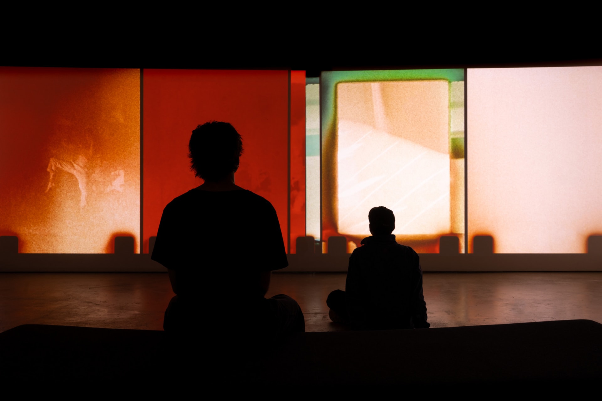 two figures sitting in front of an abstract colourful film projection in a gallery setting