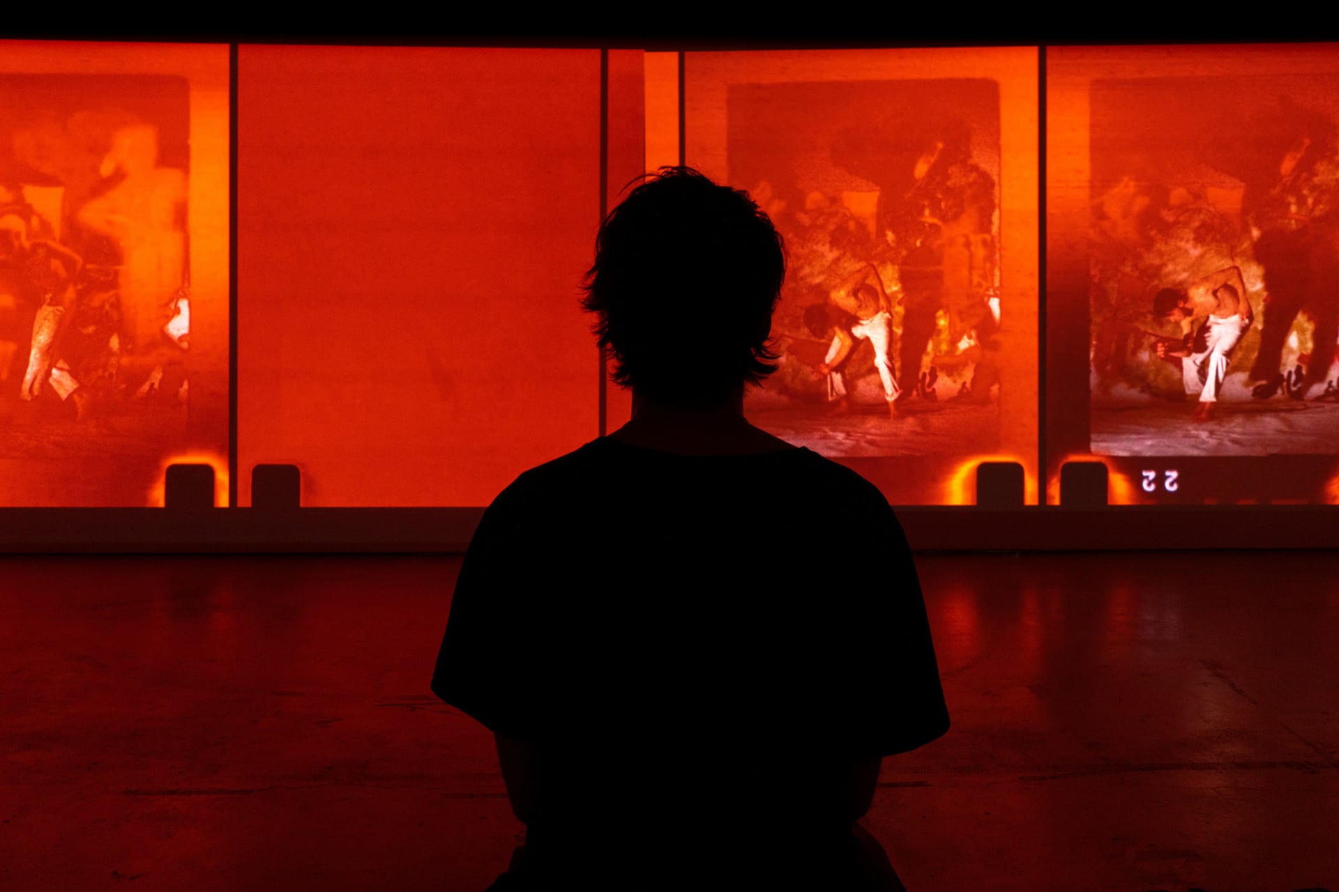 a back of a person sitting in an exhibition room watching a film projection consisting of red film light leaks