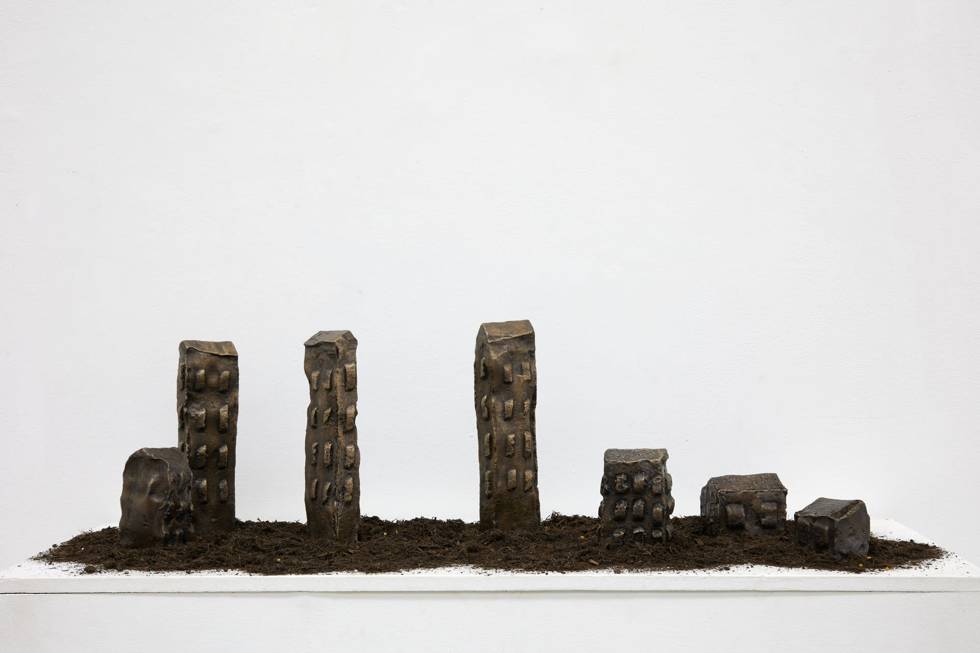 Seven Bronze disfigured towers standing side by side on a plinth.