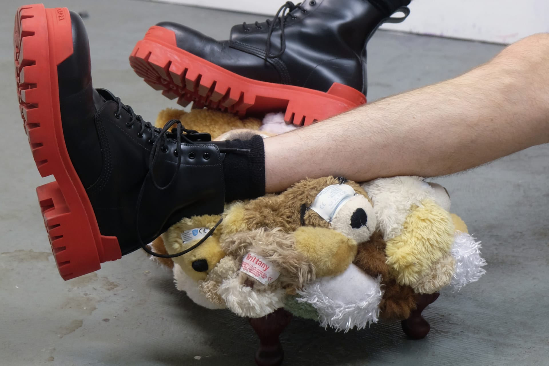 A person wearing red boots rests their feet on a footstool upholstered with soft toy body parts.