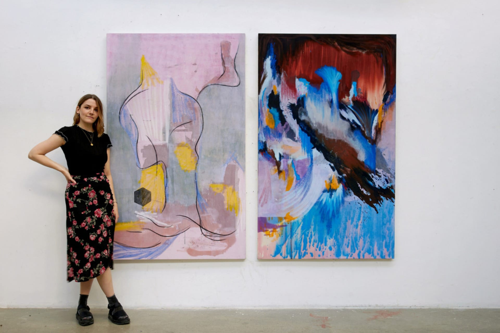 Artist standing left of 2 paintings: left is pink with green and yellow shapes, charcoal lines; right is blue with brown splash.