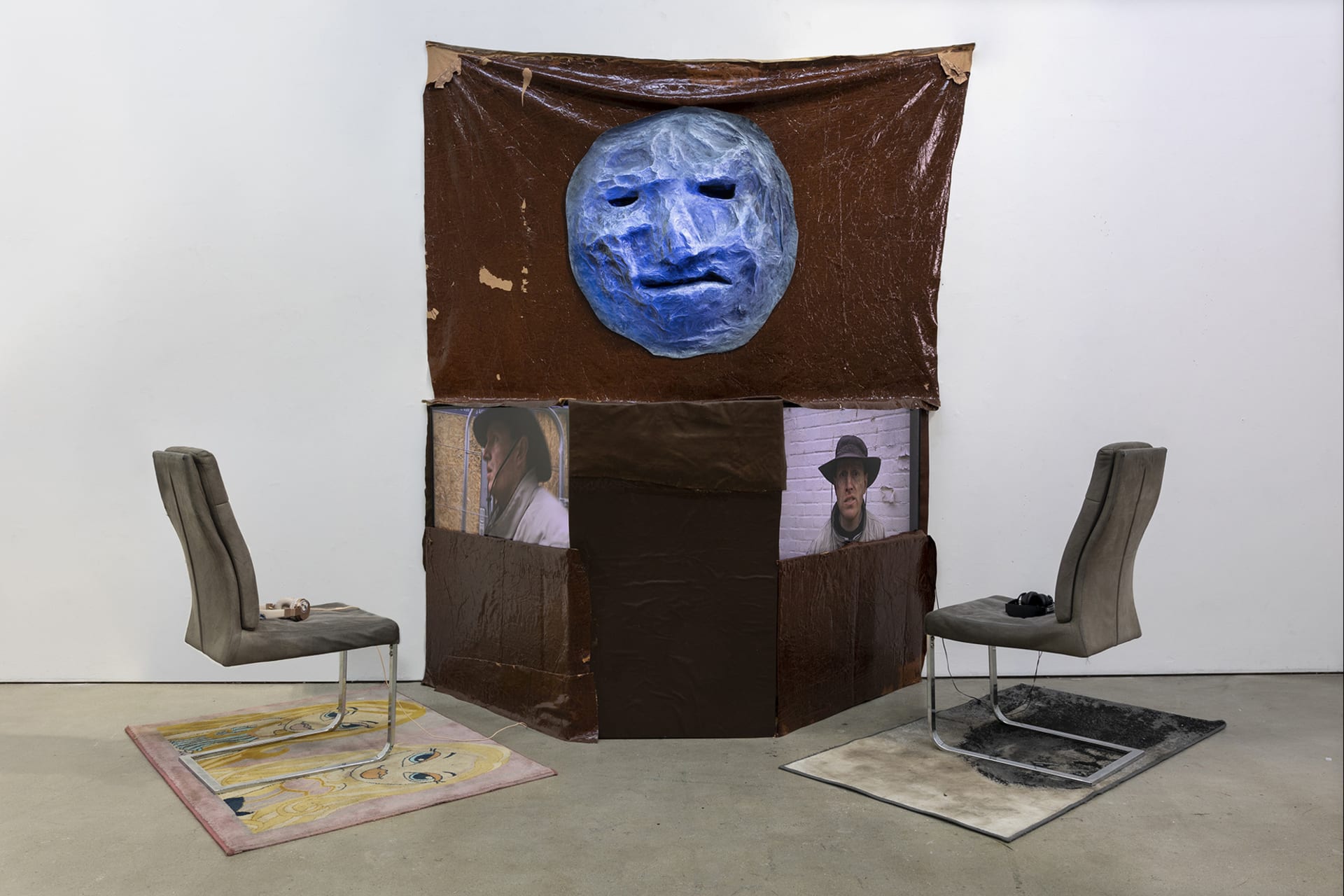 A sculpture  of a man's face hangs above large structure which houses two screens. rugs and chairs face the screens.