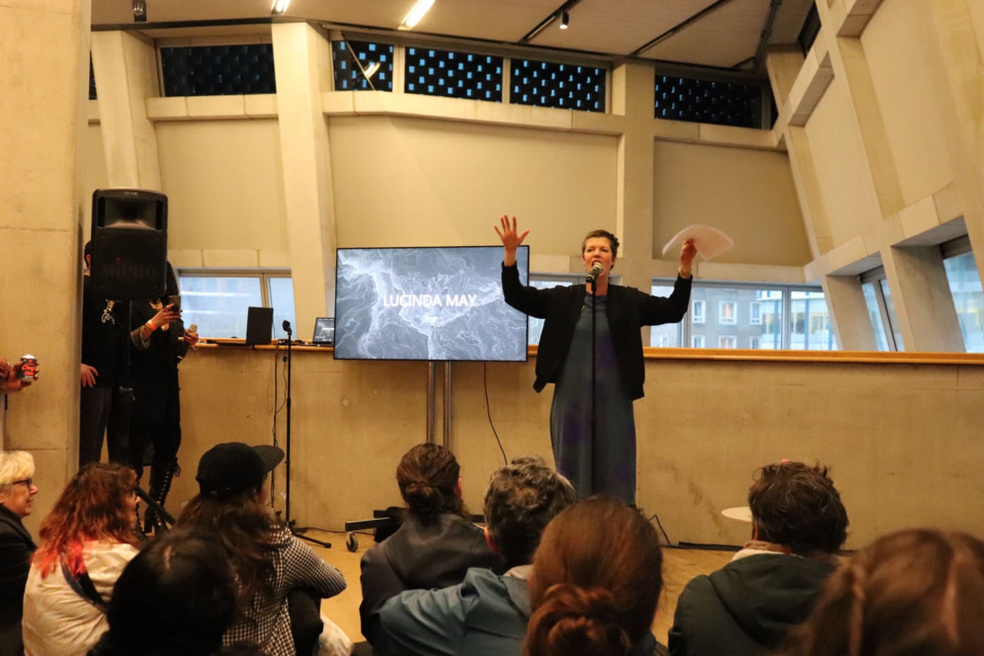 A woman sings into a microphone at Tate Modern with her hands raised. People sit on the floor in front of her.