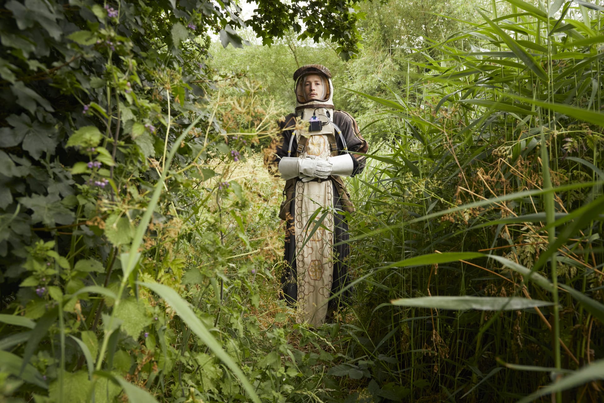 A person dressed in a costume resembling a medieval monk astronaut. They are surrounded by dense foliage.
