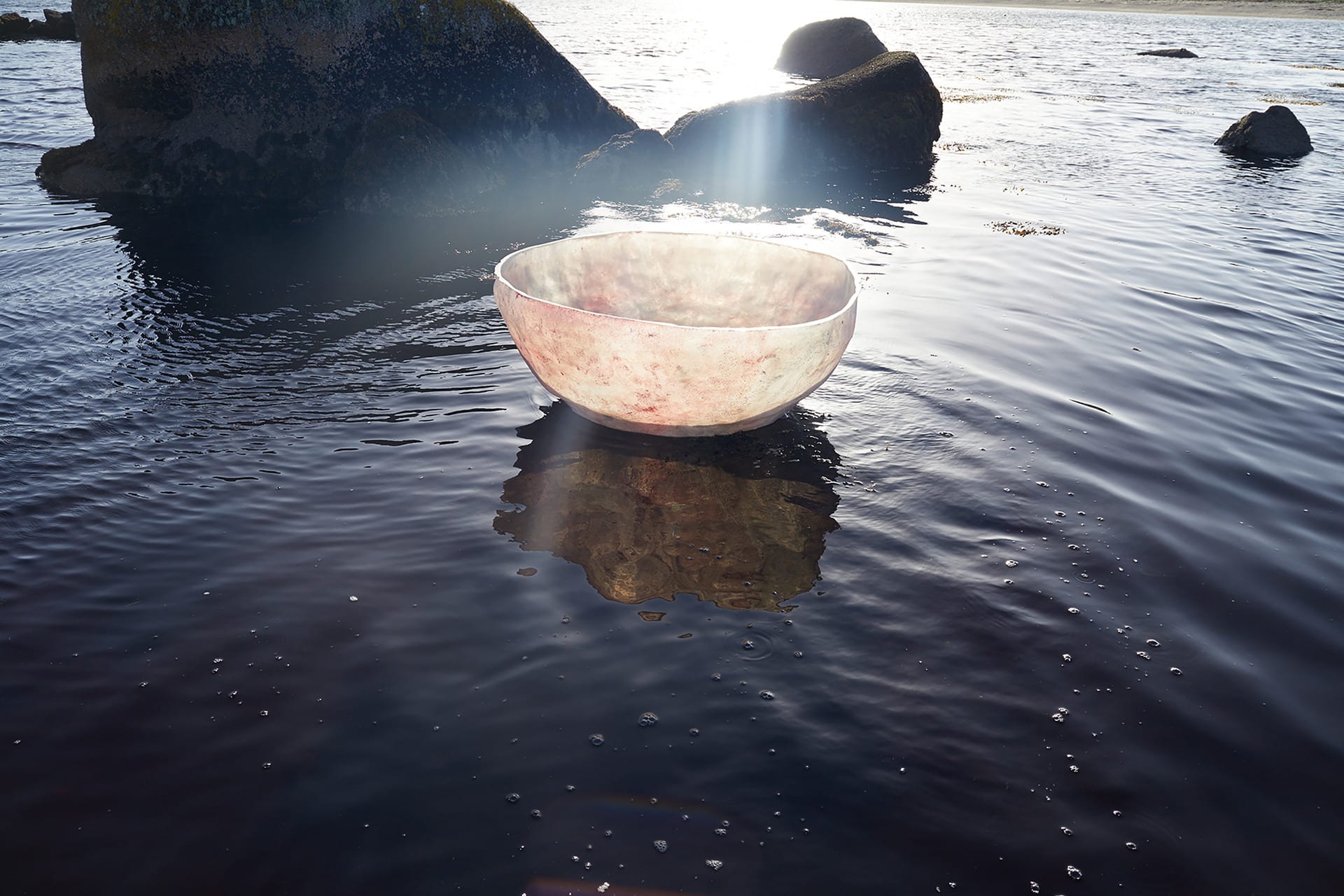 A backlit pink coracle floats on dark water in front of looming rocks