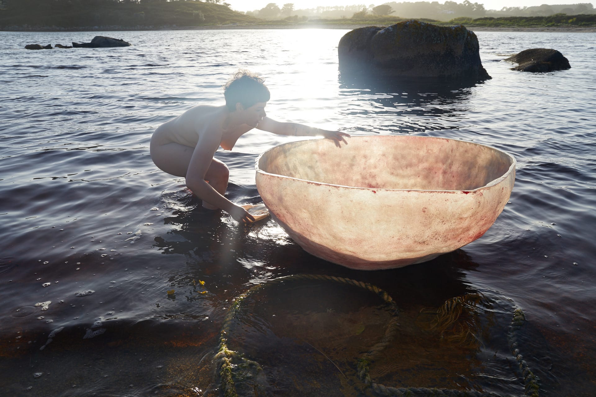 A naked person floats beside a backlit fibreglass coracle at twilight