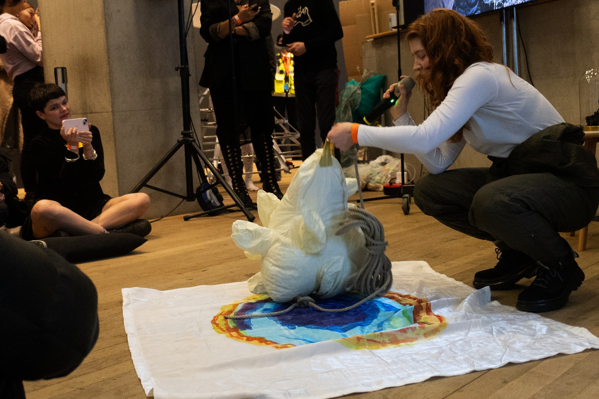 The artist performs to an audience at the Tate Modern, with a film playing on a screen in the background. They carry props.