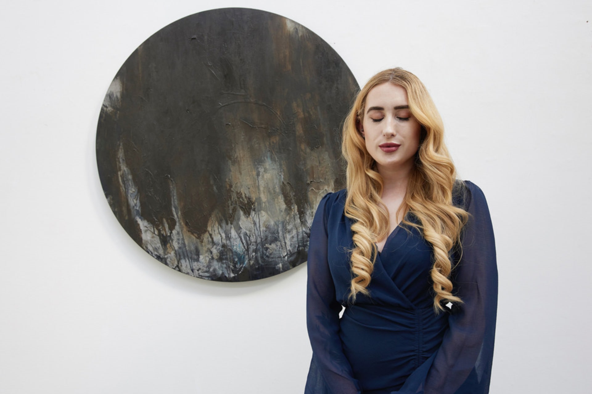 Kimberley stands in front of a large, round abstract canvas. It is brown and grey. Kimberley is a white woman with blonde hair.