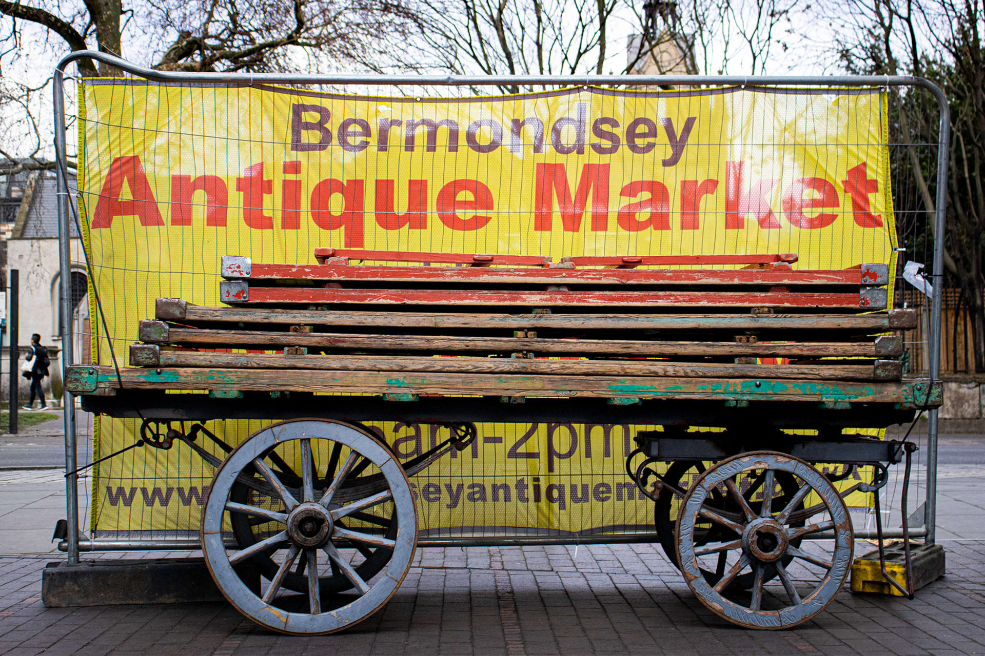 A sign and very old and large cart with planks on it rests on cobblestones. The sign reads boldly "Bermondsey Antiques Market"