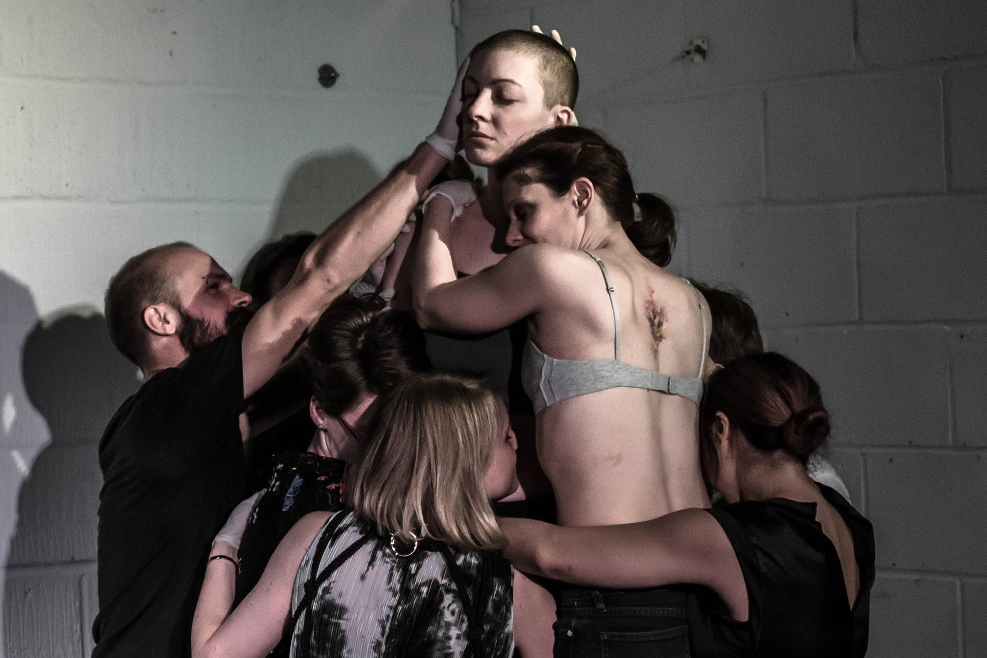 Shaved-head female performer in black shirt and pants, standing on a crate. A group of spectators hugging her. 