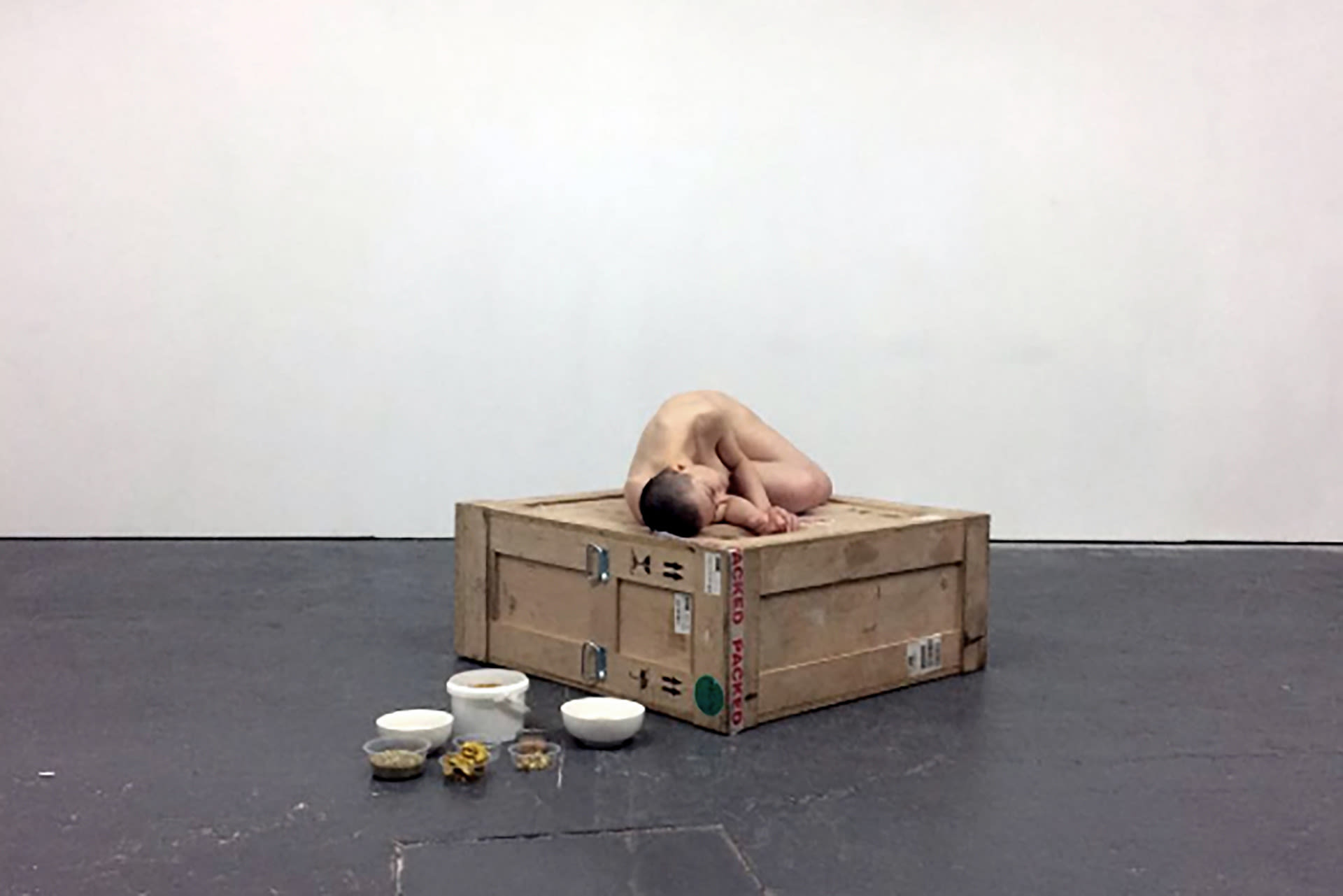 Shaved-head female performer lying naked on a crate with bowls of glitter and honey placed on the floor right next to her. 