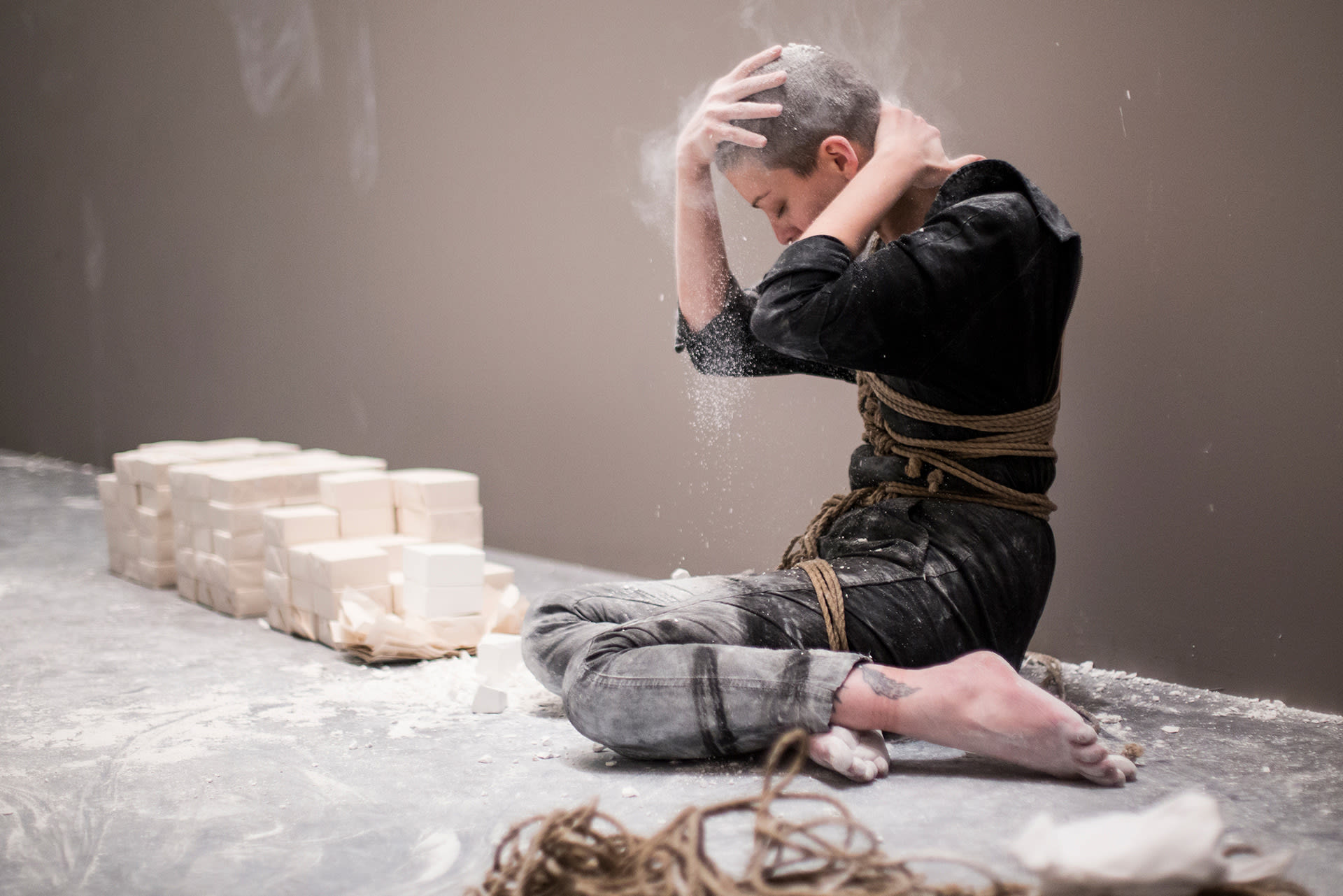 Shaved-head female performer in a worker's uniform, in bondage, sitting on the floor, covering her head with chalk. 