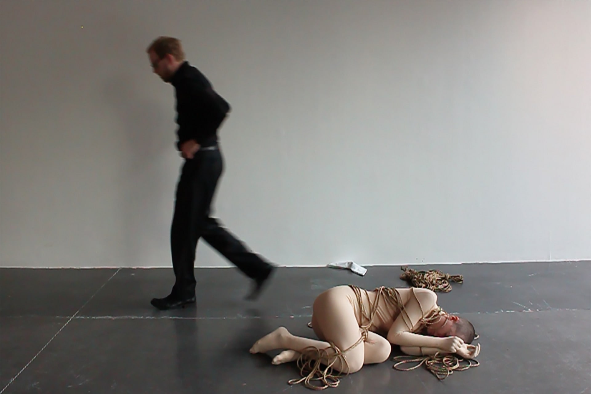 Shave-head female performer, wearing a skin-toned full body-suit, lying on the floor and being abandoned by a male rigger.