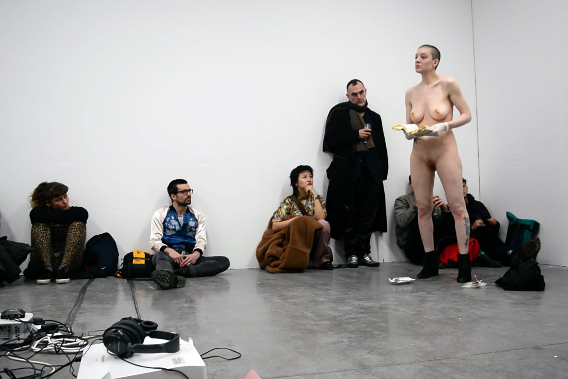 Shaved-head, naked, female performer, standing up and reading from a book. 