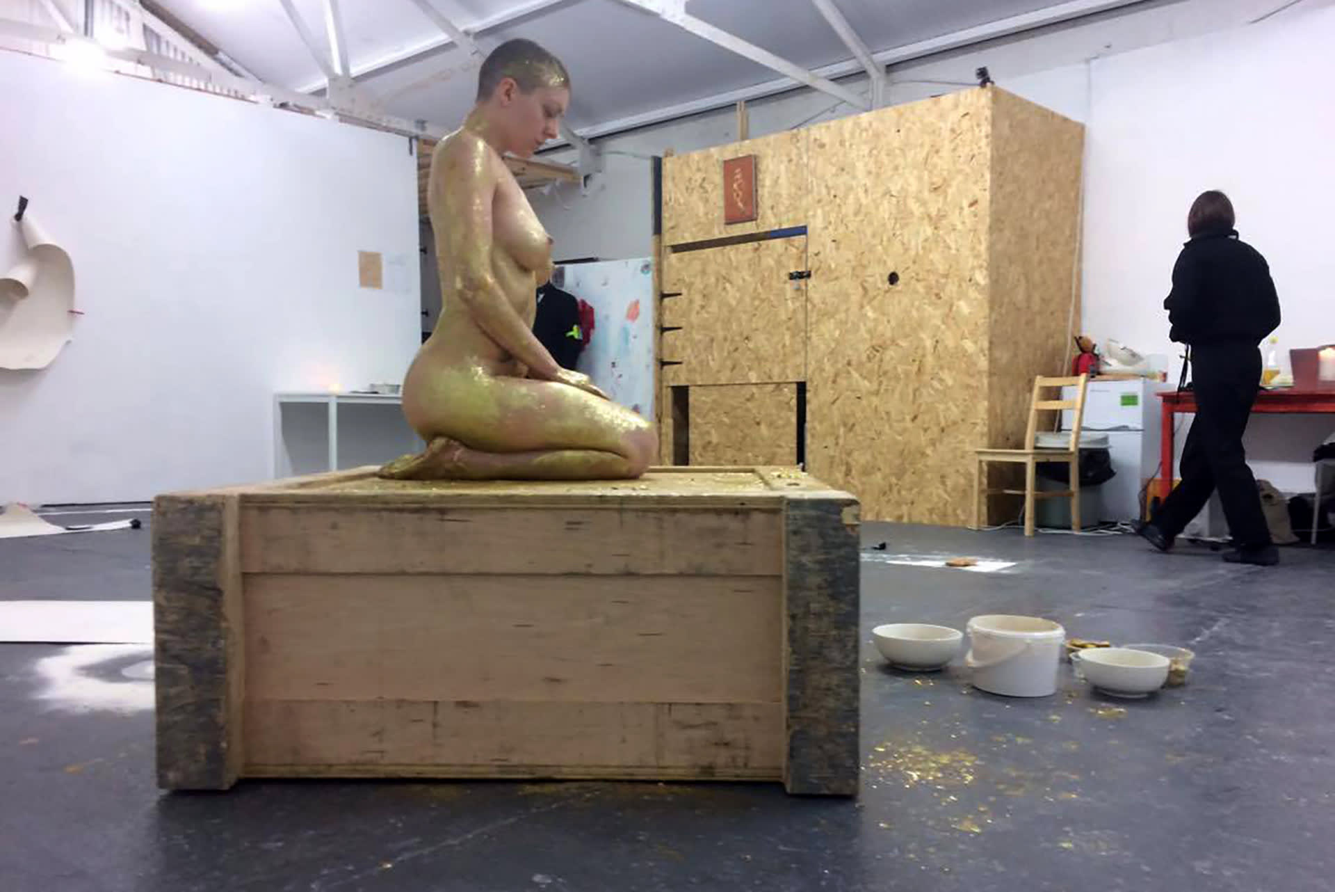 Shaved-head female performer kneeling on top of a crate, with her body covered in glitter and honey. 