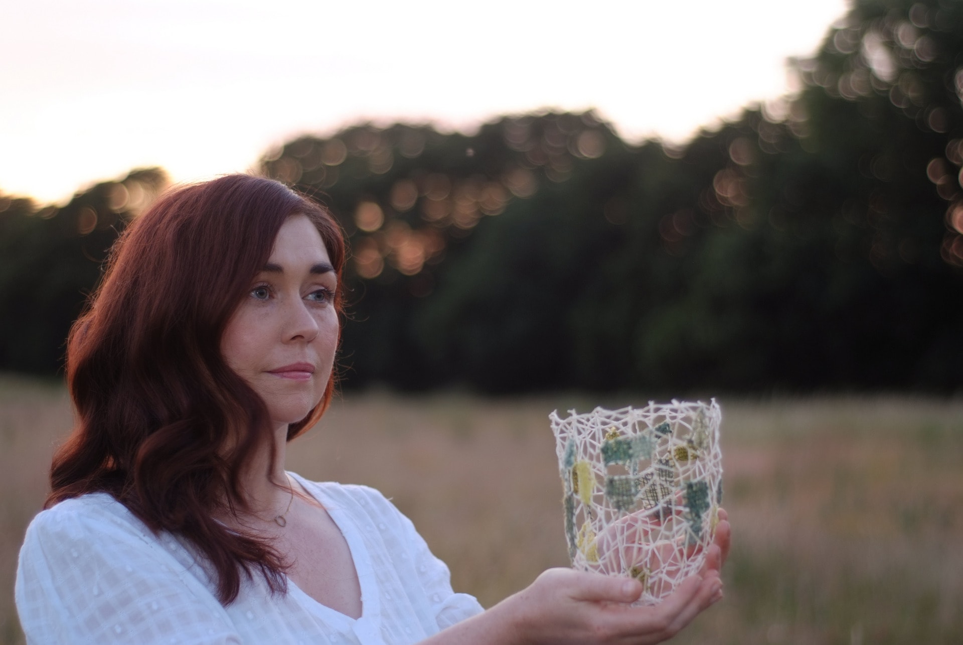 Joanne holds a white and green vessel outside in a spring meadow. She is wearing a white top and has auburn hair. 