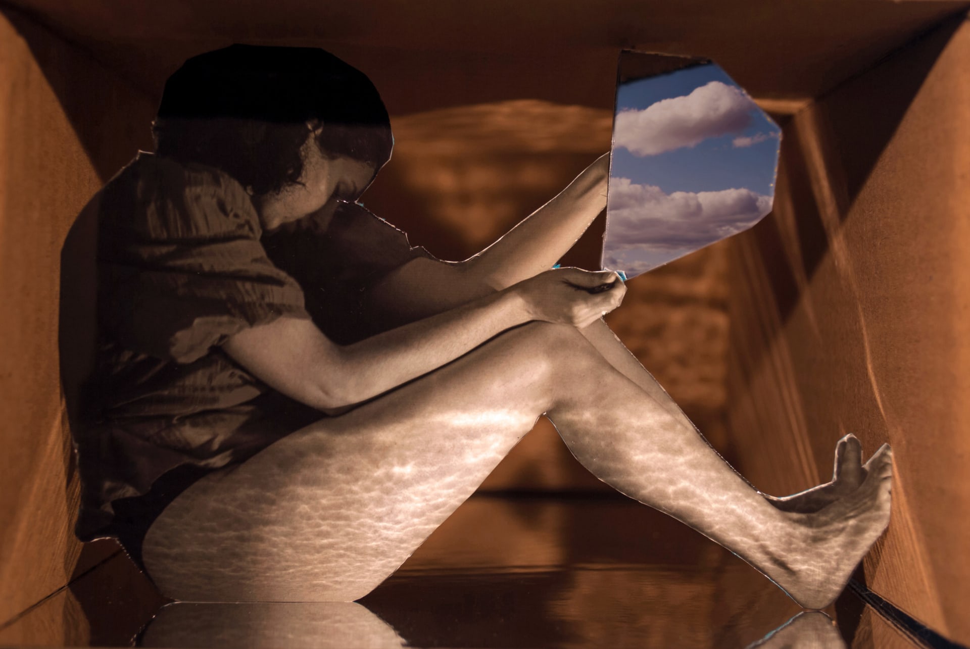 Side view of a cardboard box containing a cut out figurine of a woman sitting and holding a mirror at the sky.