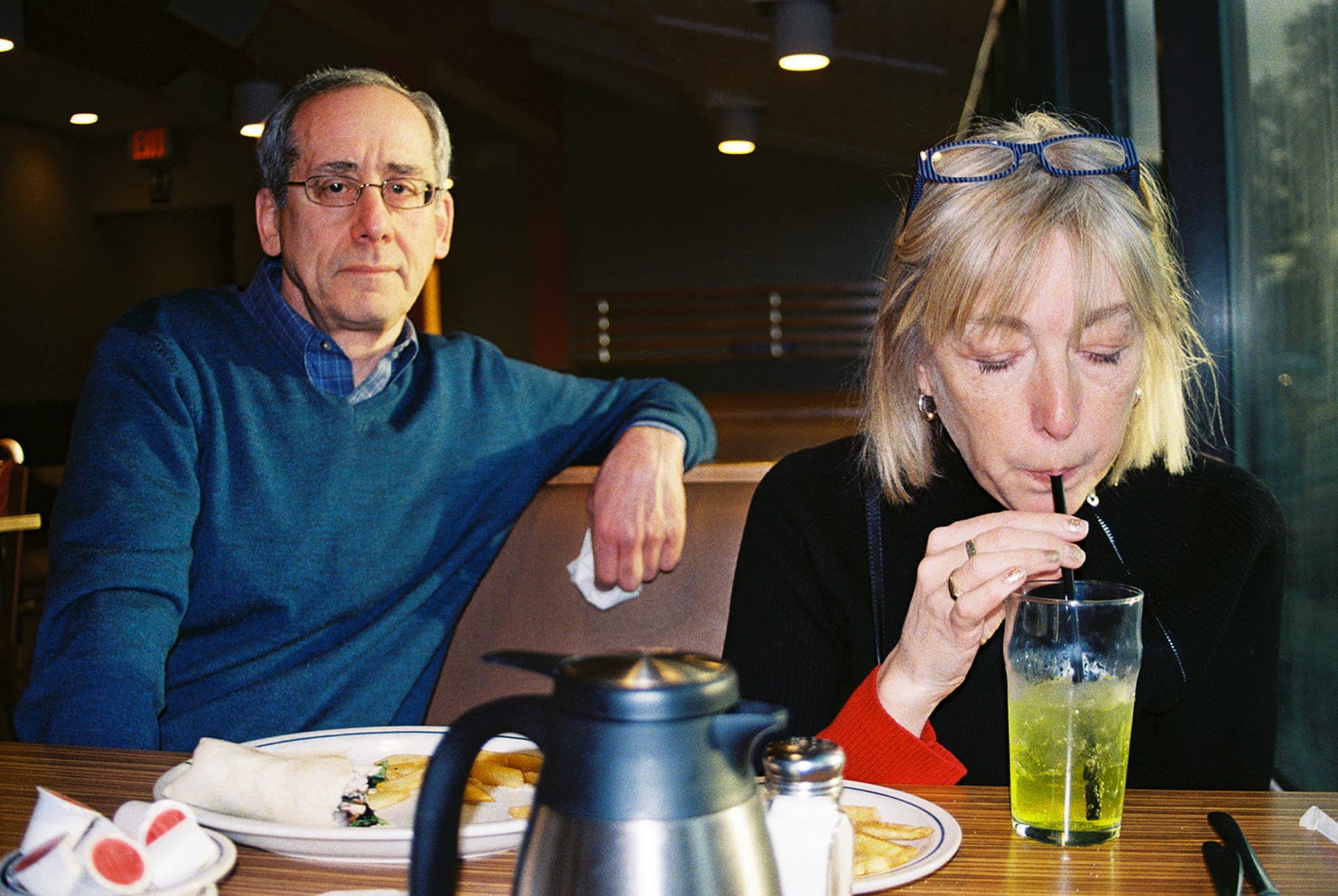 Man and Woman sitting side by side in a restaurant dining booth. The man looks at the viewer. Woman is sipping a drink.