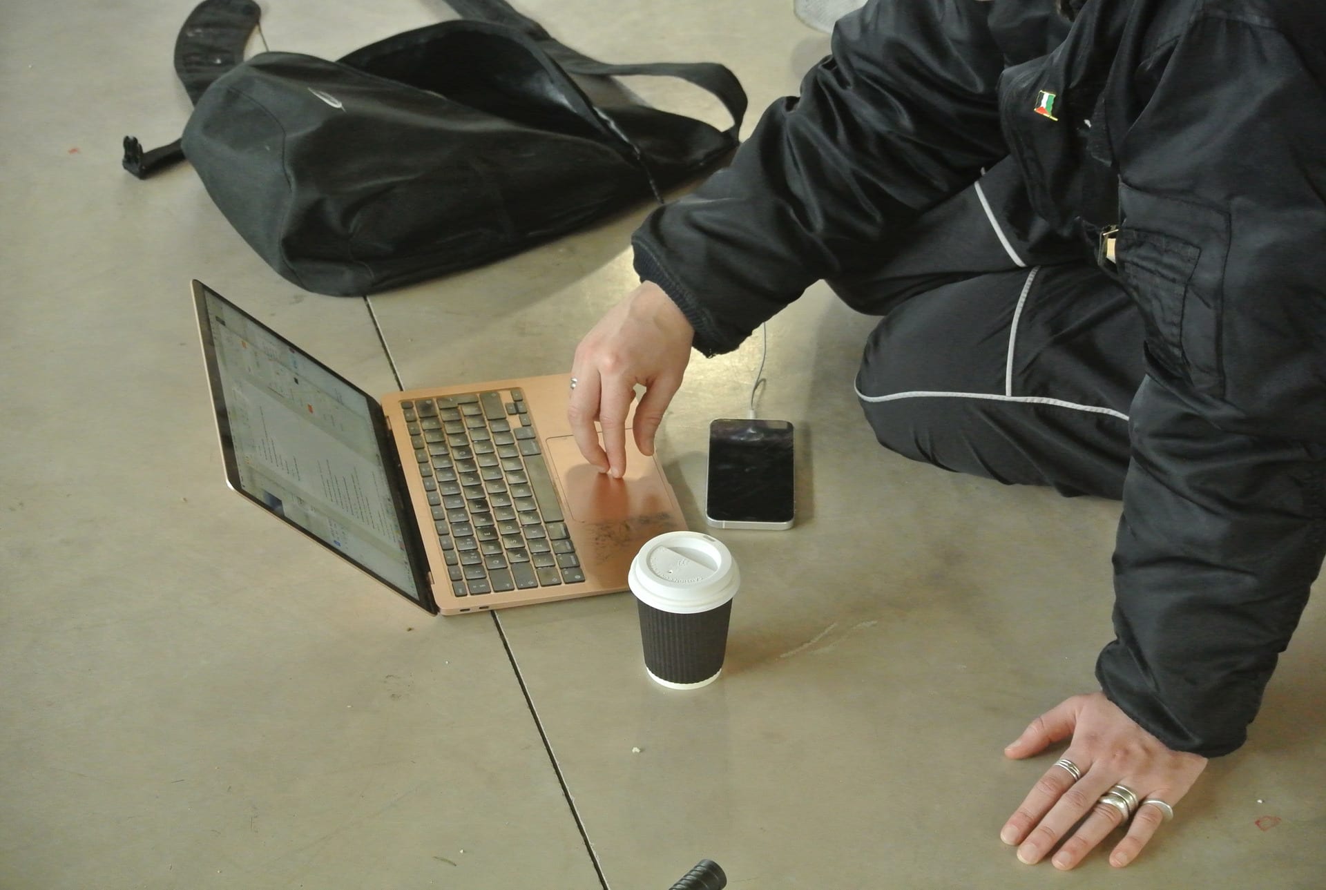 a person sits on the floor and is typing on their laptop. a bag, a coffee cup, and a phone are on the floor. 