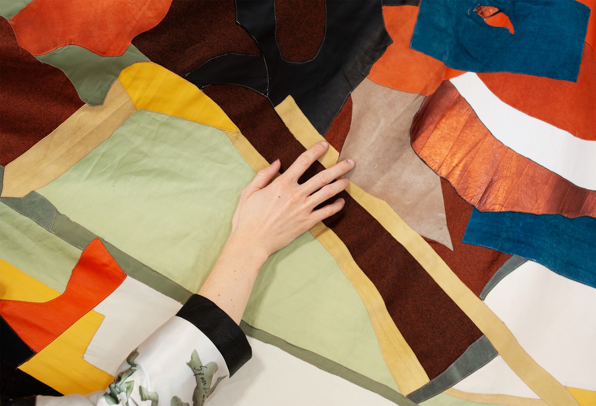 A hand is patching different colored fabircs and leather together to form a giant patchwork. 