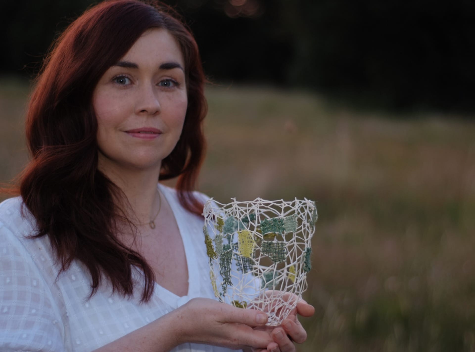 Joanne stands holding one her of handwoven baskets outside in a meadow. She is wearing a white top and has auburn hair. 