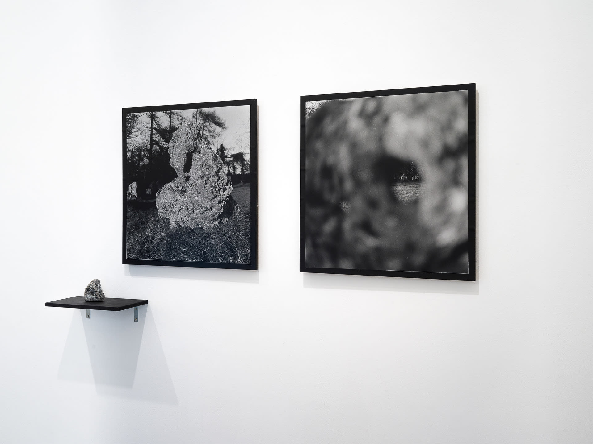 Two black and white photographs on a white wall, showing the close up details of a standing stone. A small shelf sits underneath