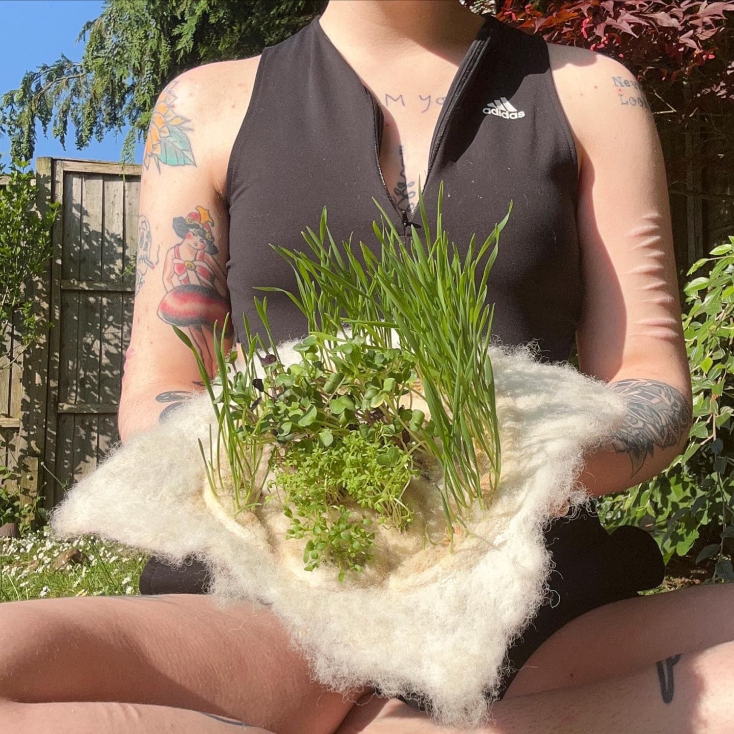  white tattooed person sitting in a garden hold a piece of white felt with several types of plants growing on it