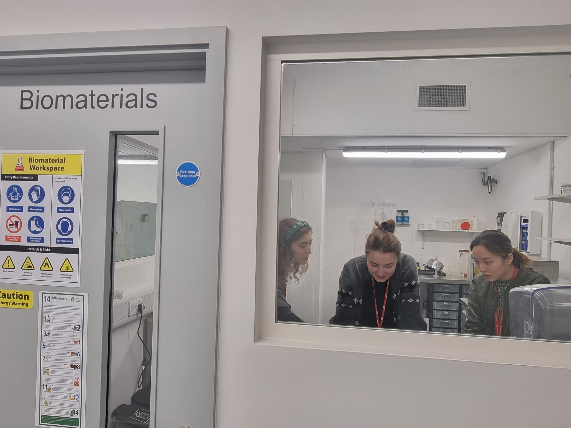 Looking through window into bio-materials lab, three team members discussing project.