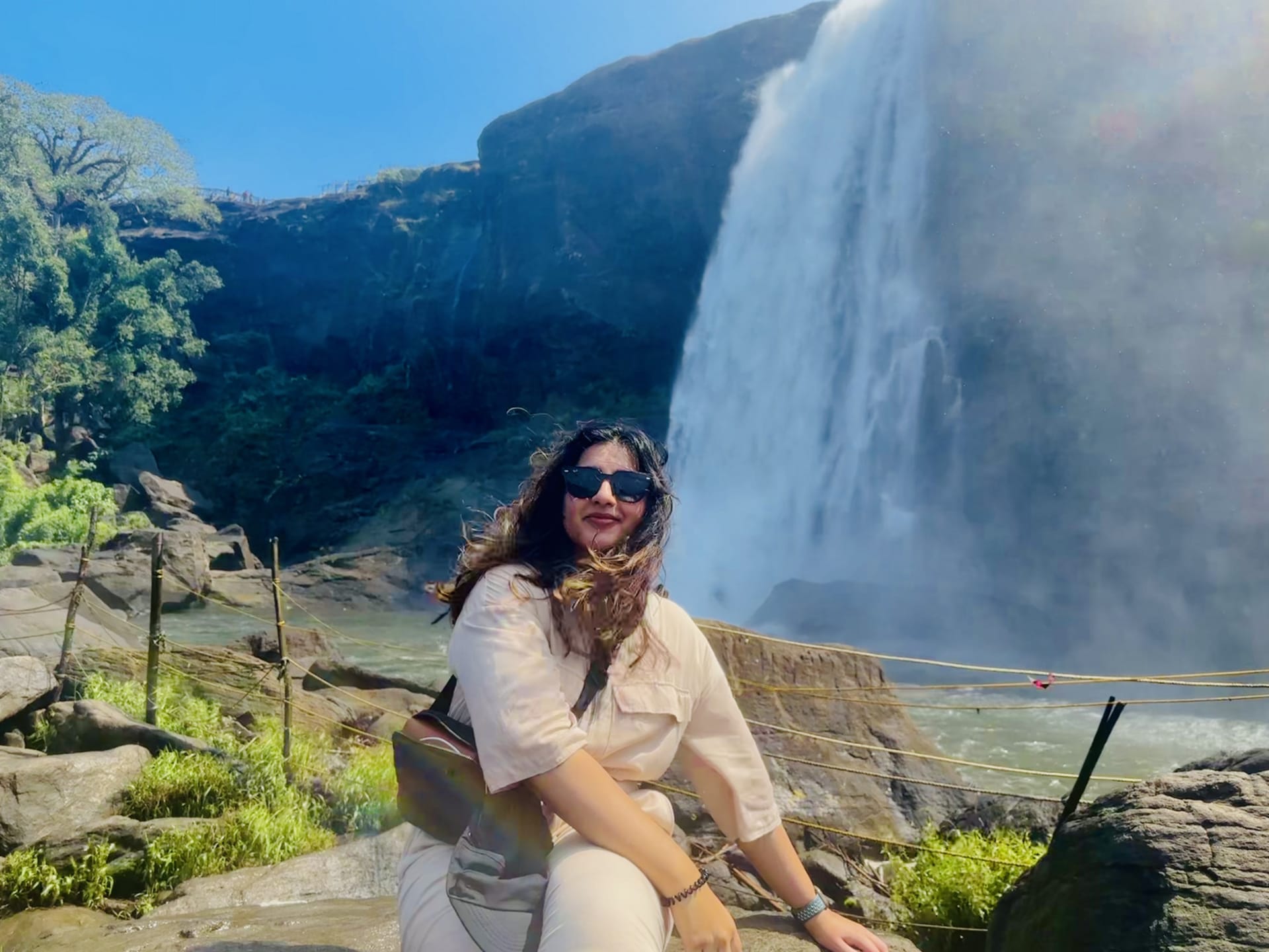 A woman is sitting on a rock while a waterfall is seen in the background.