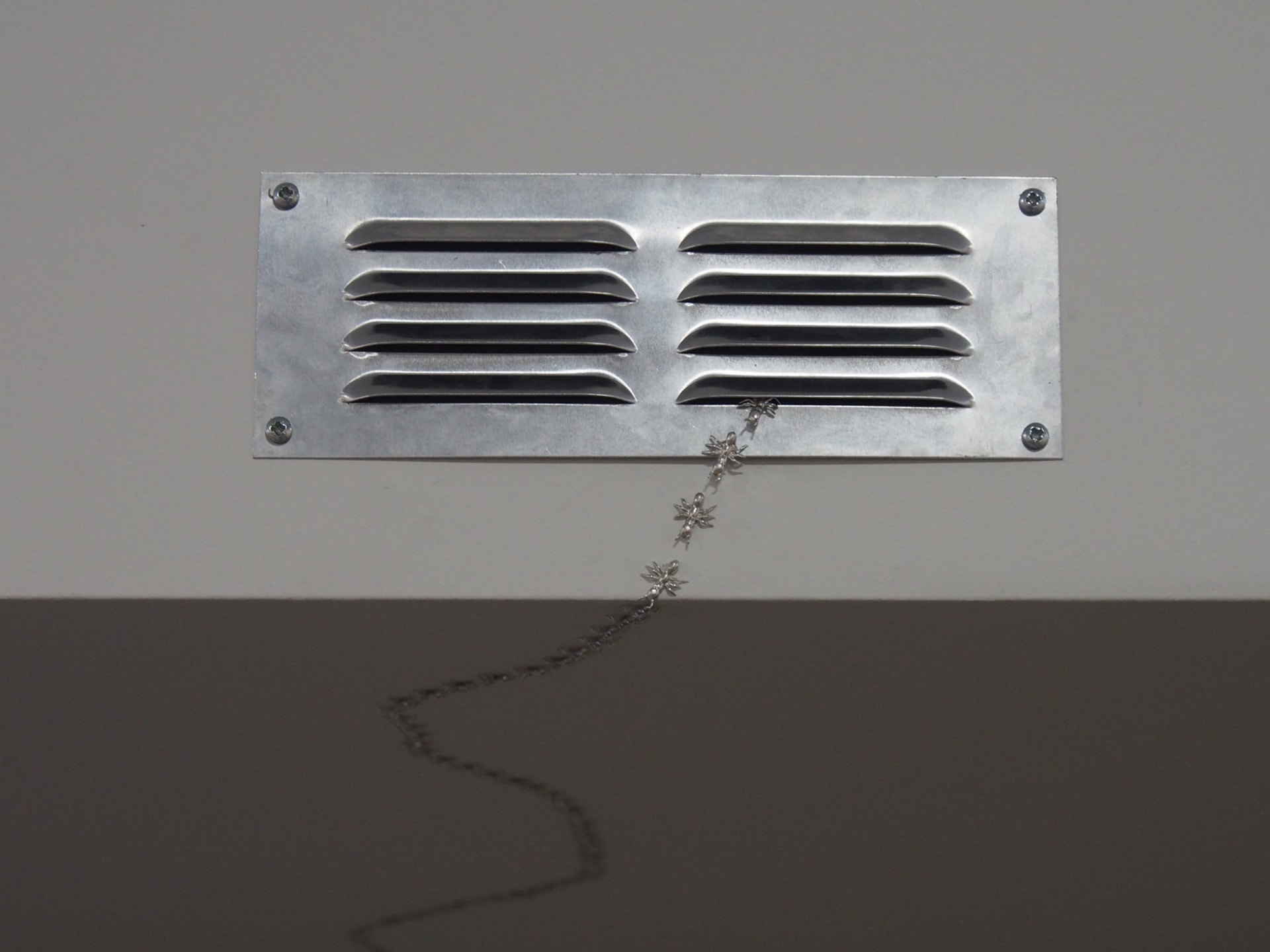 A silver vent attached to a white wall with a trail of polished silver ants coming from it.