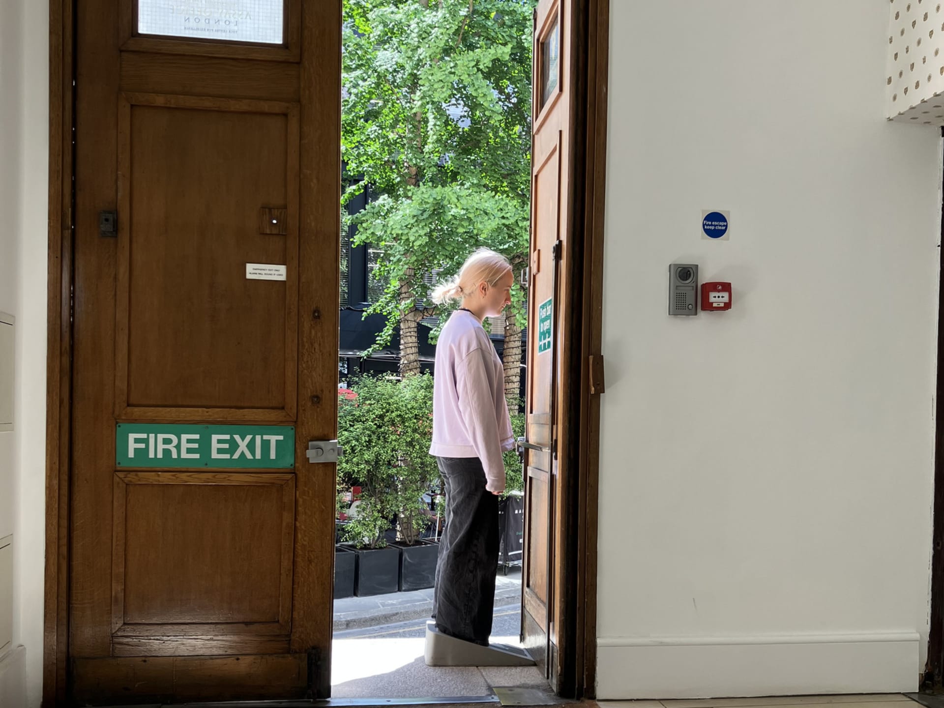 Lili is wearing a large doorstop shaped grey rubber show and is holding the door open with it at the london assay office