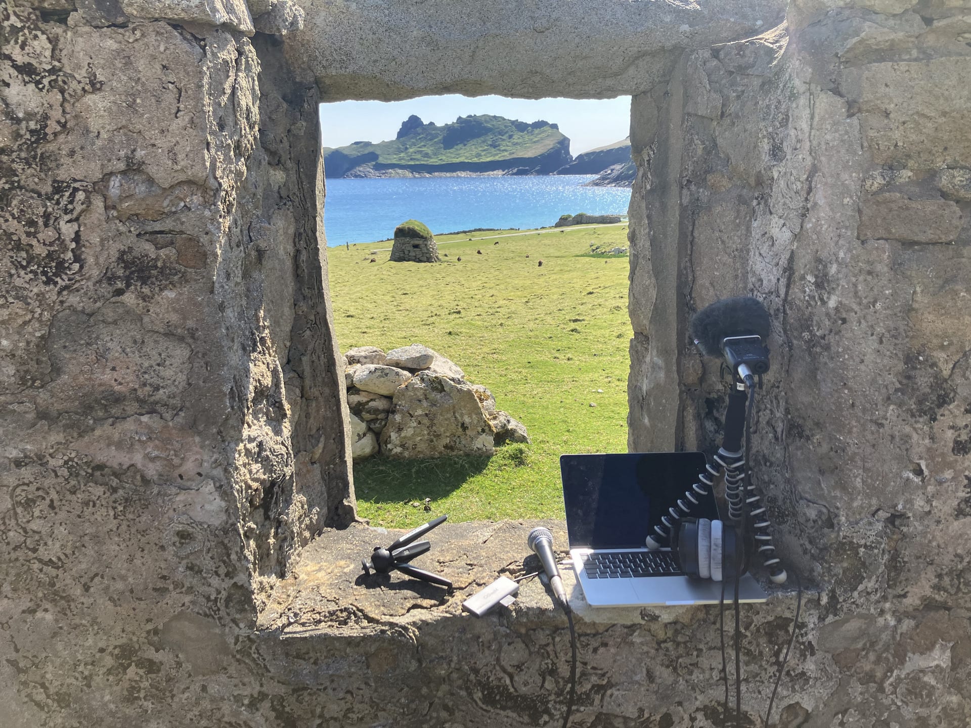 A laptop and microphones sit on a sunny old stone croft window sill without glass. The view through is grass, sea, cliffs, sky.