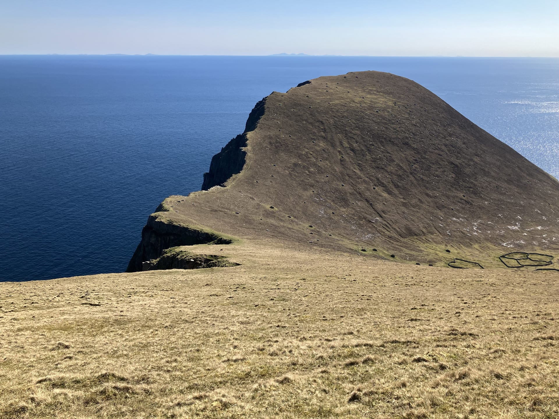 A hill cliff rises abruptly out of blue sea. The view is from above it with sea all around. On the right the hill slopes down.