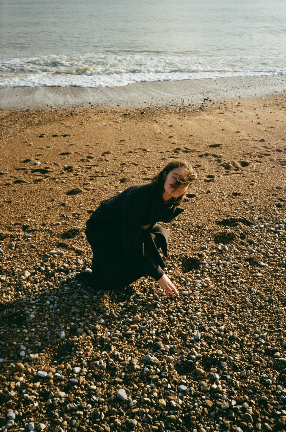 35mm photograph of me on Hastings beach in a long dark grey coat. I am crouching down to pick up a stone, my face obscured