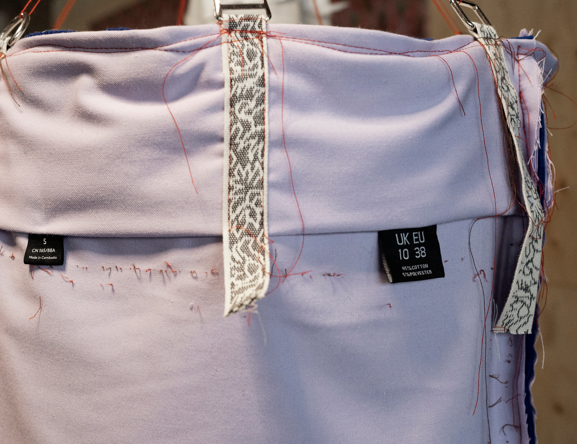 Close-up detail of lilac fabric with chains attached by ribbon, two clothing labels and many unpicked seams exposing red thread