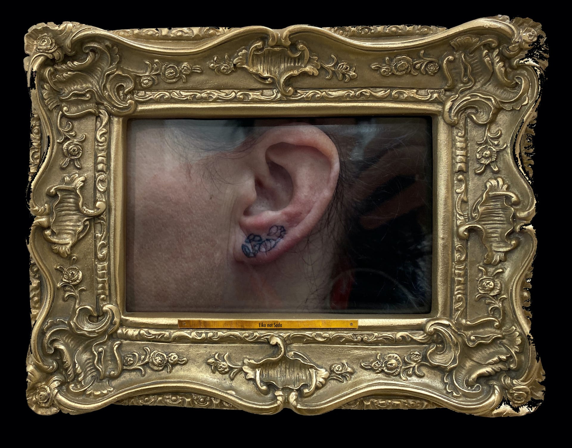 Photographic image of artists left ear with scorpion tattoo in a gold baroque frame with nameplate reading "Eiko not Sada"