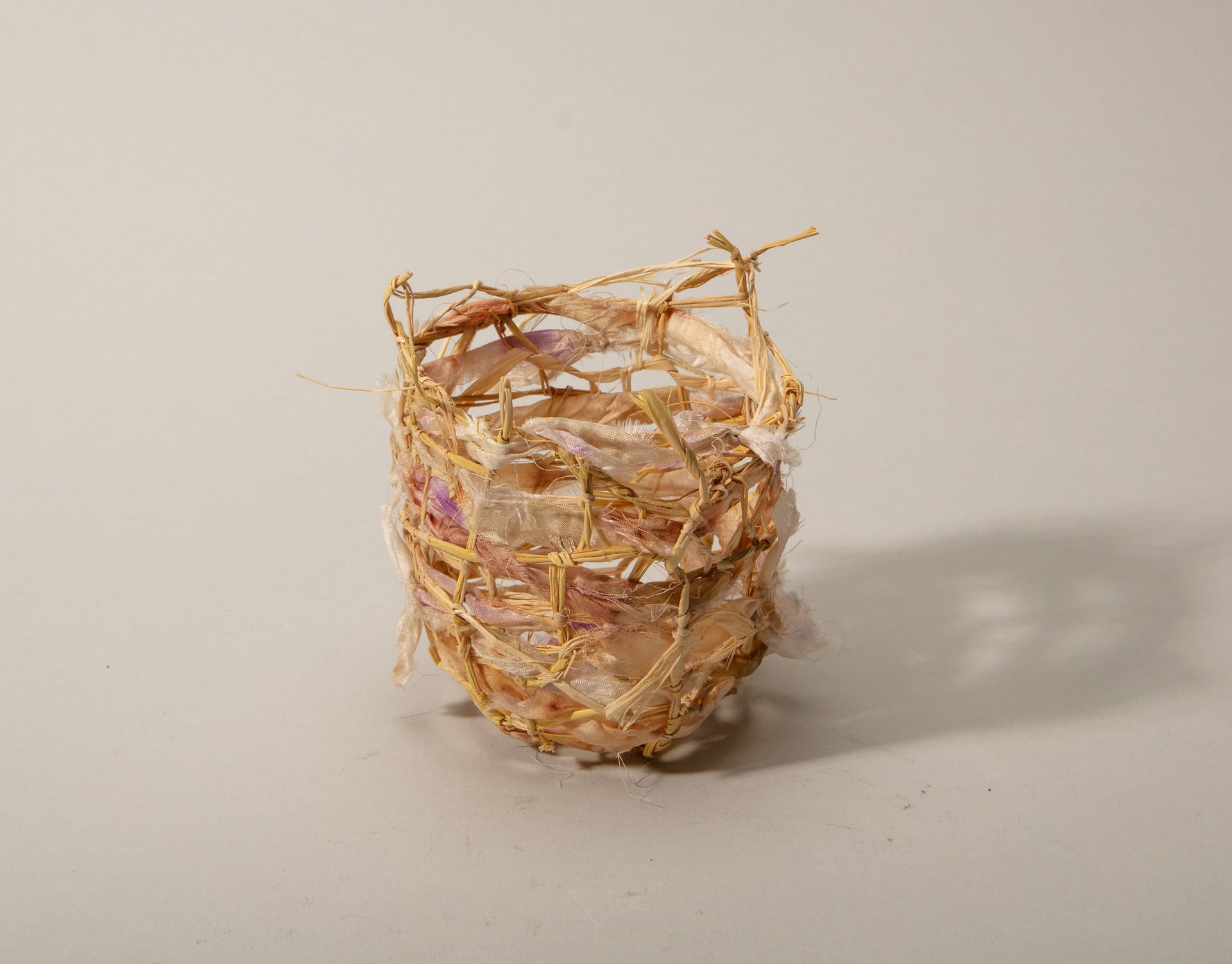 A small basket woven with raffia and bundle dyed pink silk is photographed against a white backdrop.