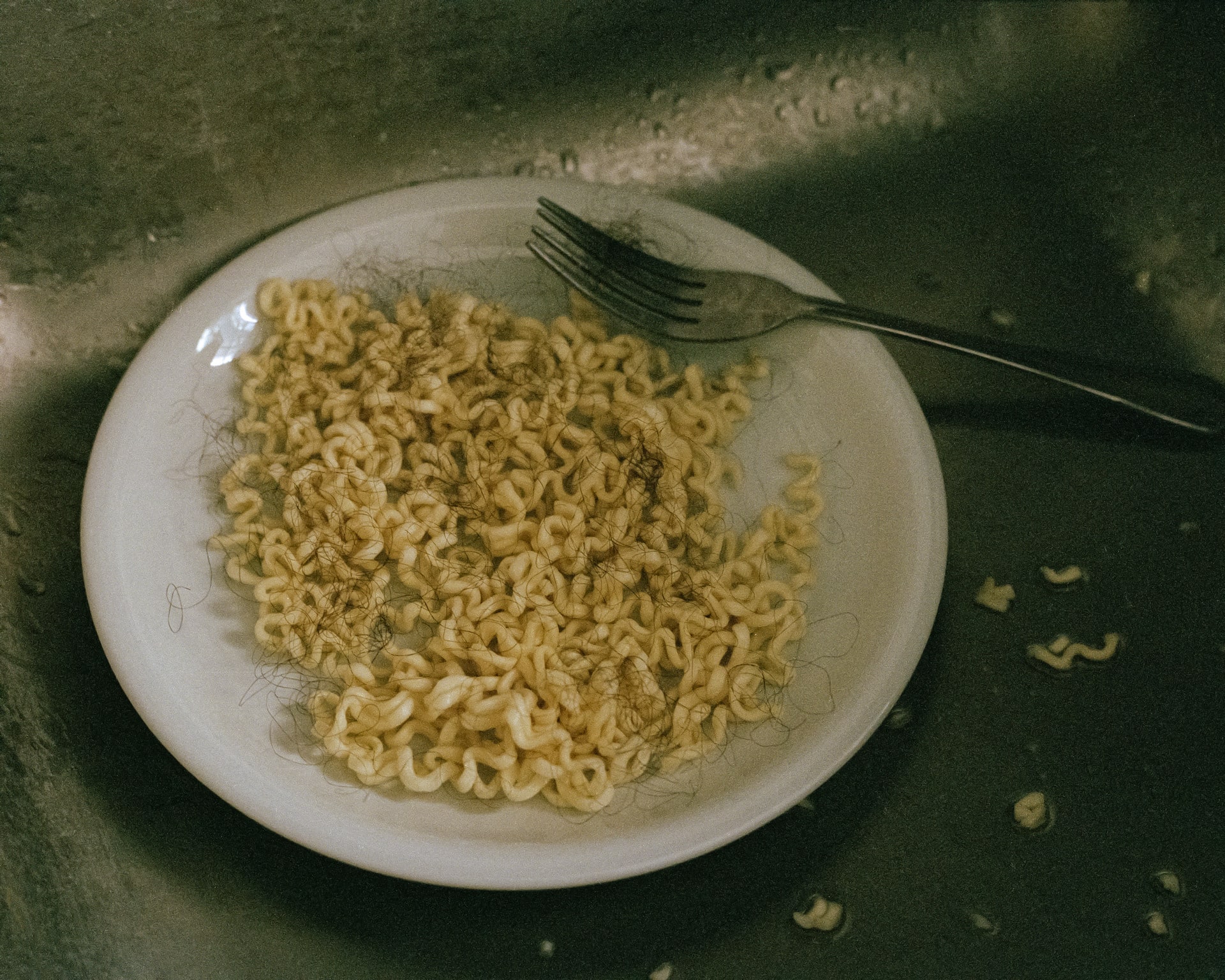 A white plate with instant noodles and curly hair is put in a silver sink.