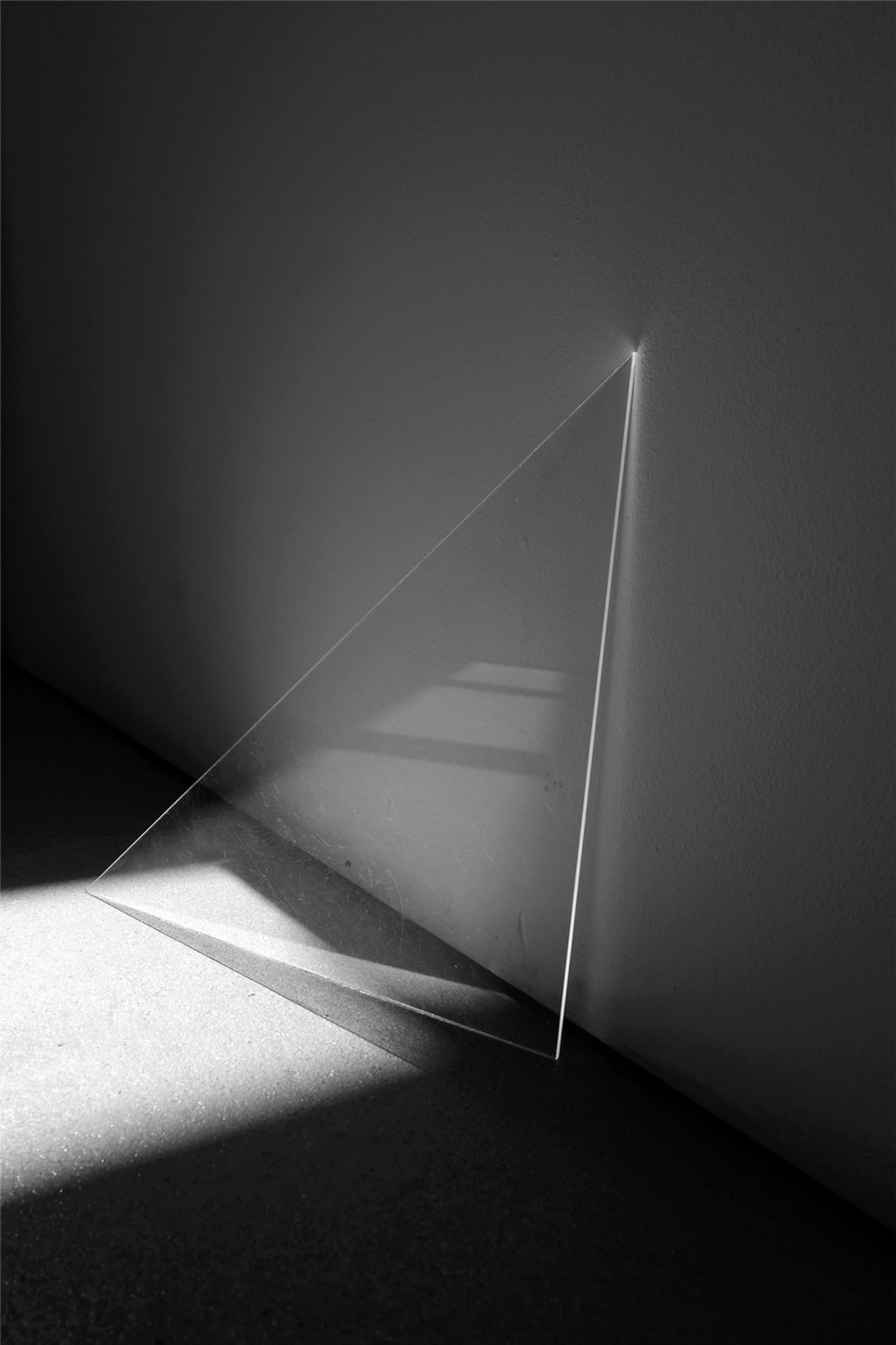 Acrylic triangle placed on the floor, leaning against a while wall. Sun light projects its highlights and shadows