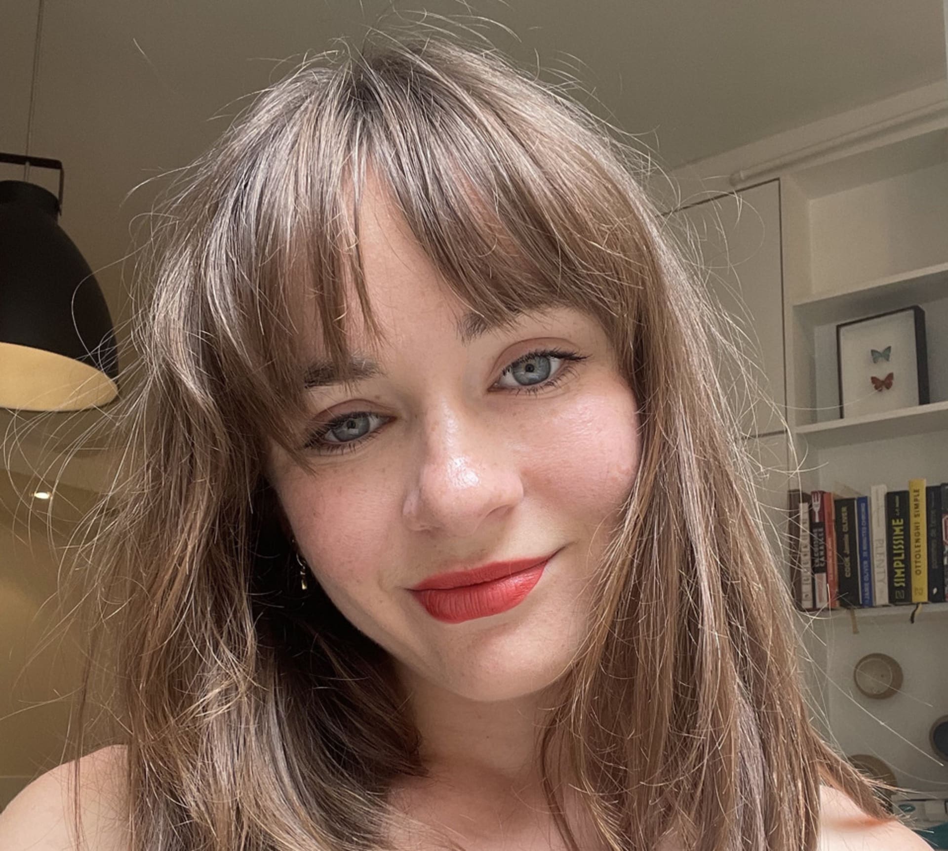 A self portrait of a woman of 23 years old smiling with brown long hair, bangs, blue eyes and red lipstick