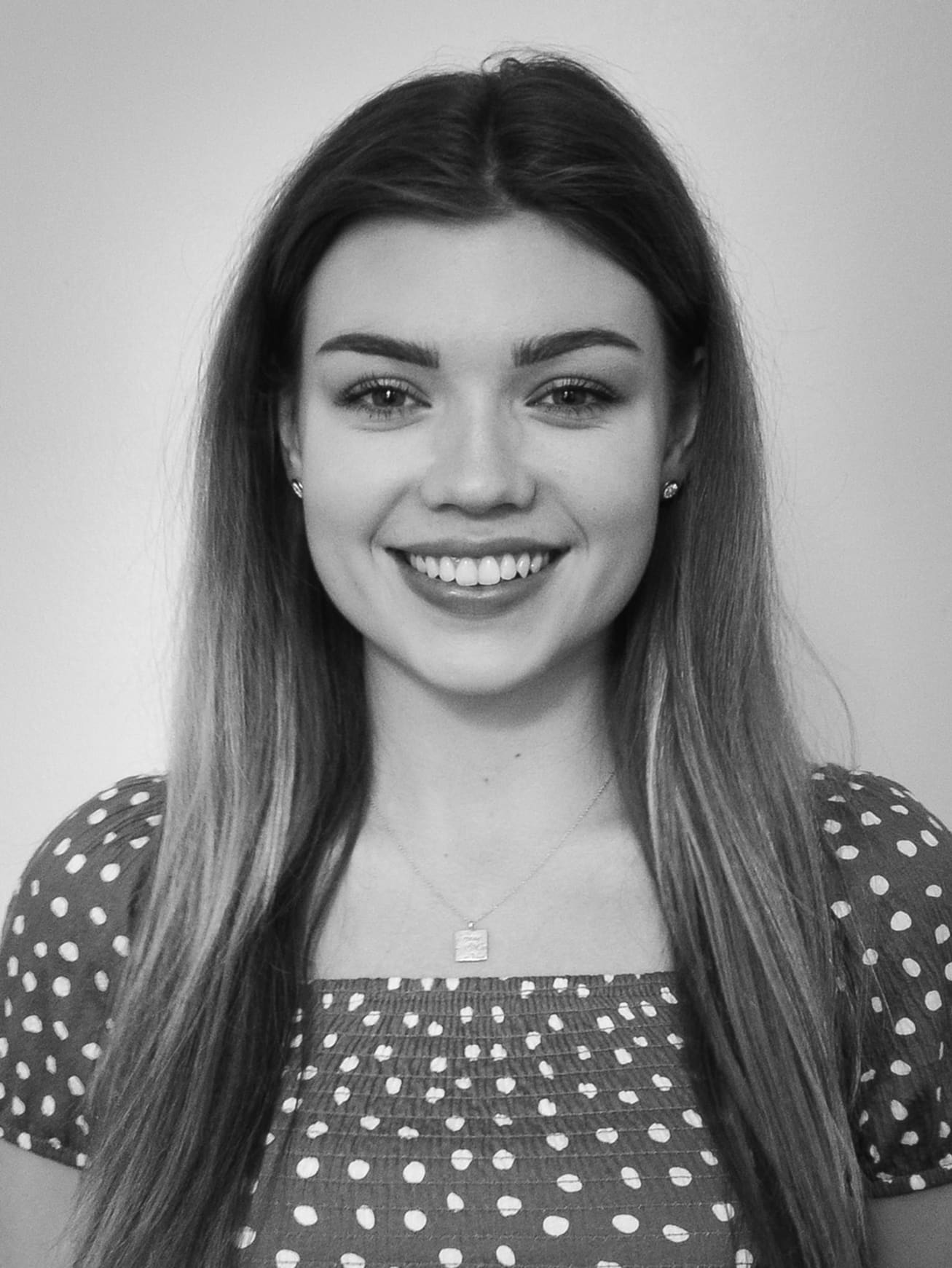 Black and white headshot of Dayna - long brunette hair, spotty blouse.