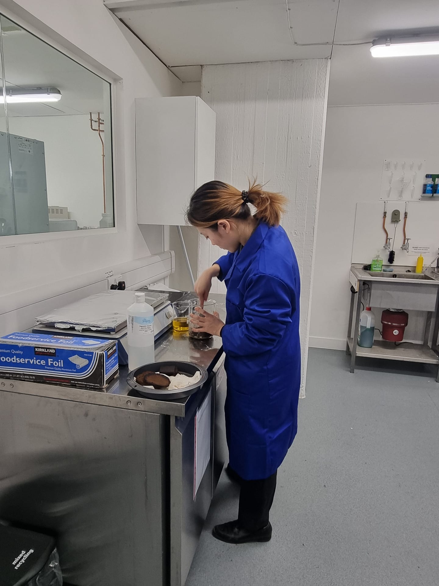 Girl in blue lab coat standing at counter in white room, mixing brown material in glass jar.