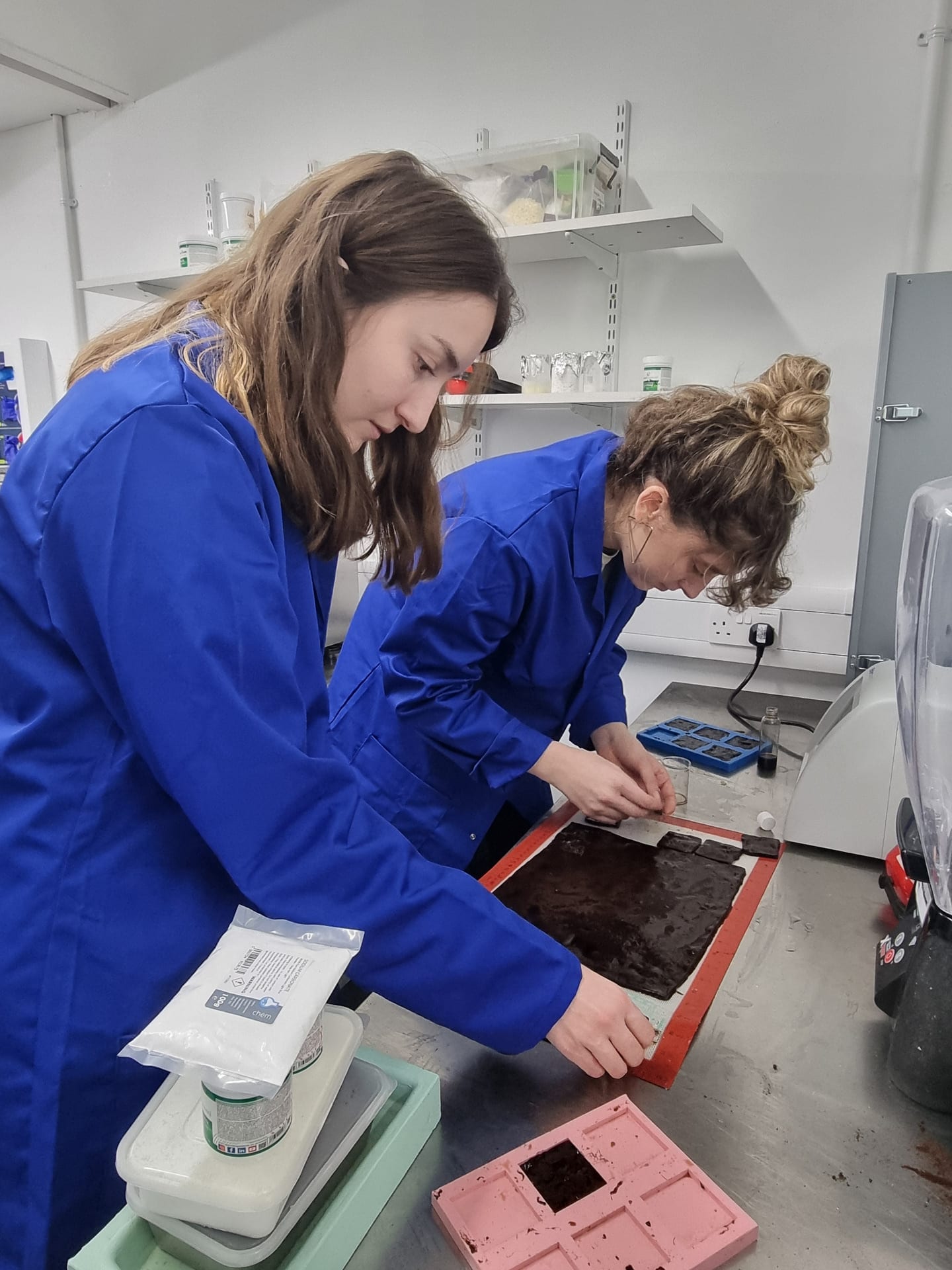 Two girls in blue lab coats standing at metal counter working on brown jelly-like material.