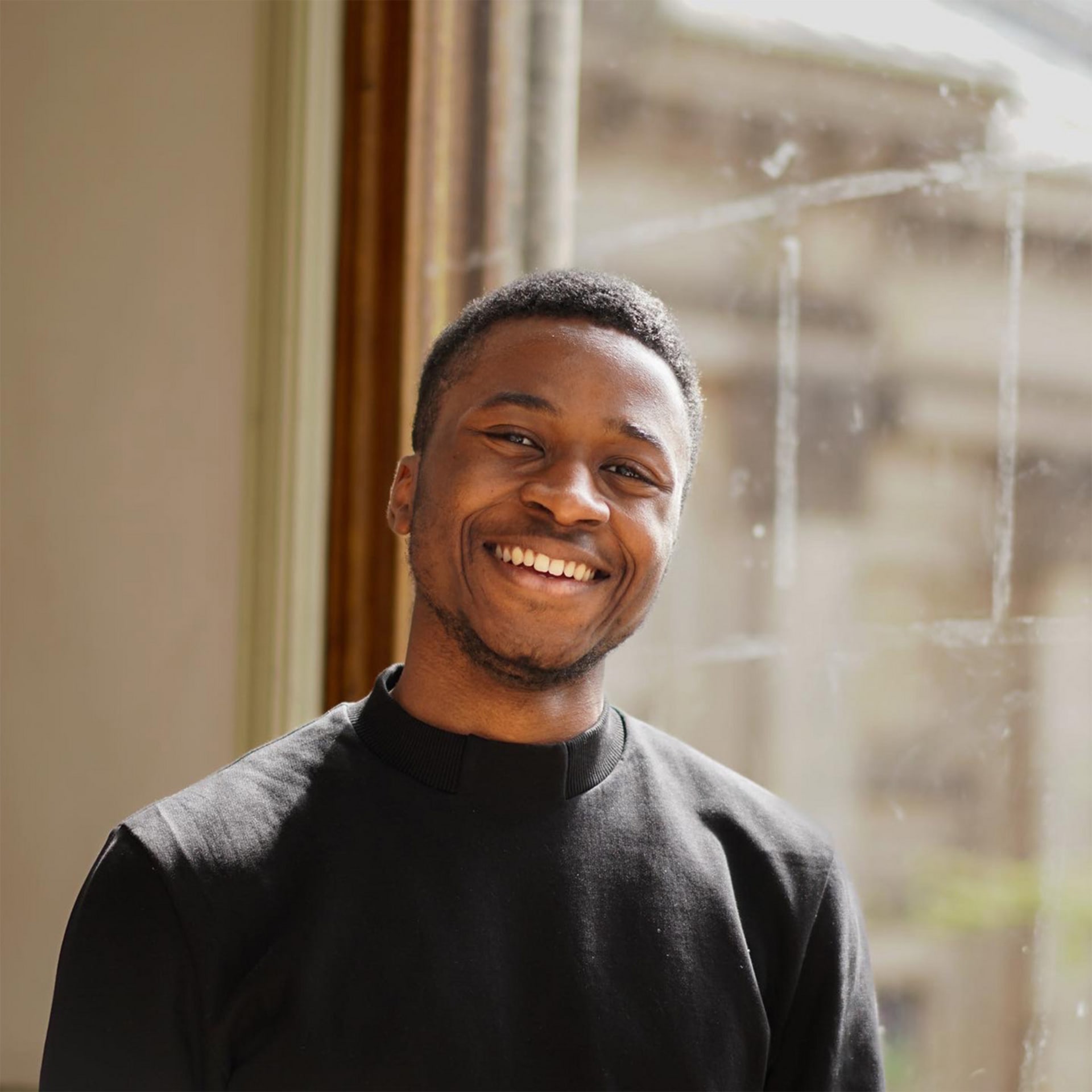 Canaan J Brown: a short-haired young Black British man, beaming at the camera, wearing a black turtleneck. He is brightly lit.