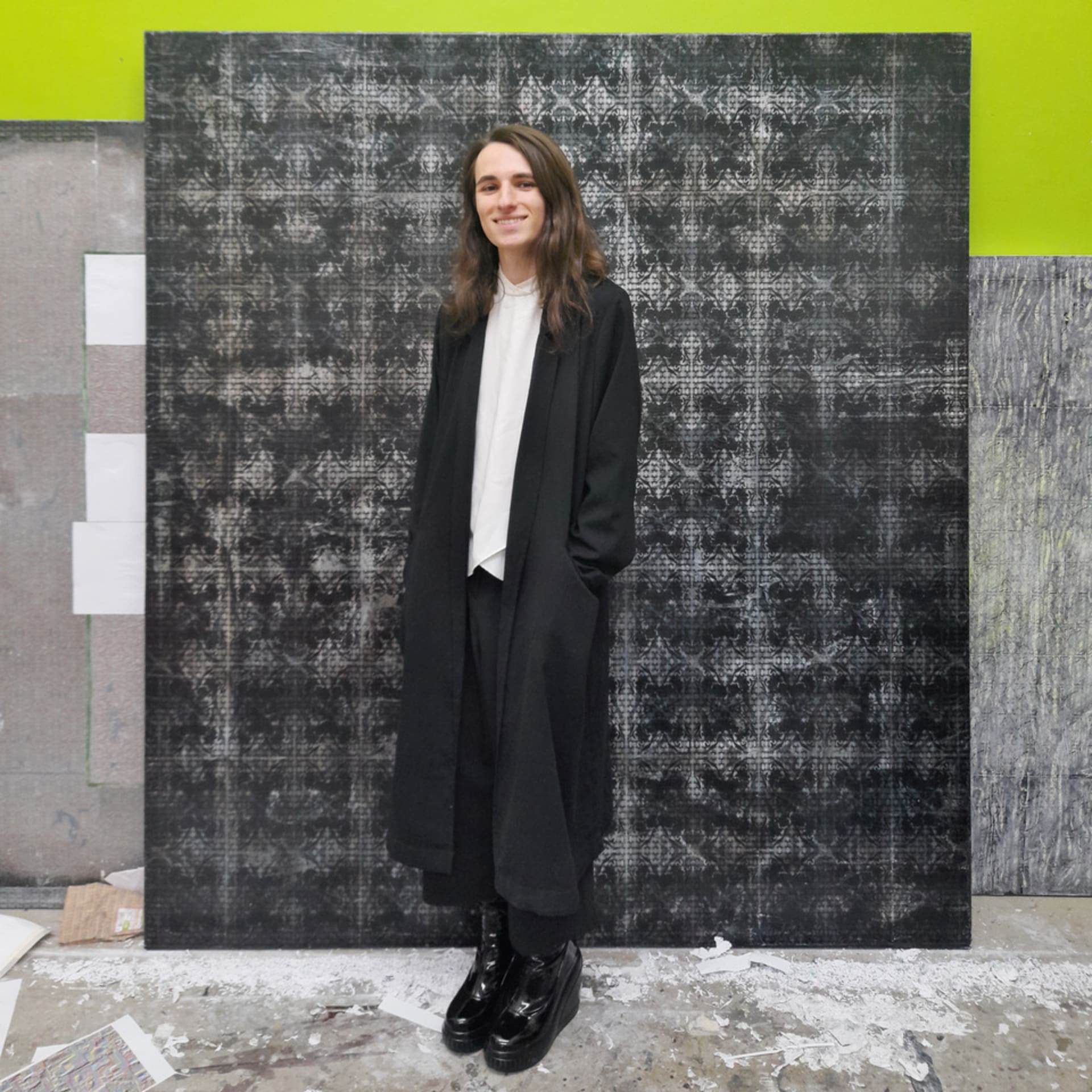 Noah Mashiak standing in front of three large paintings stacked against his bright green studio wall.