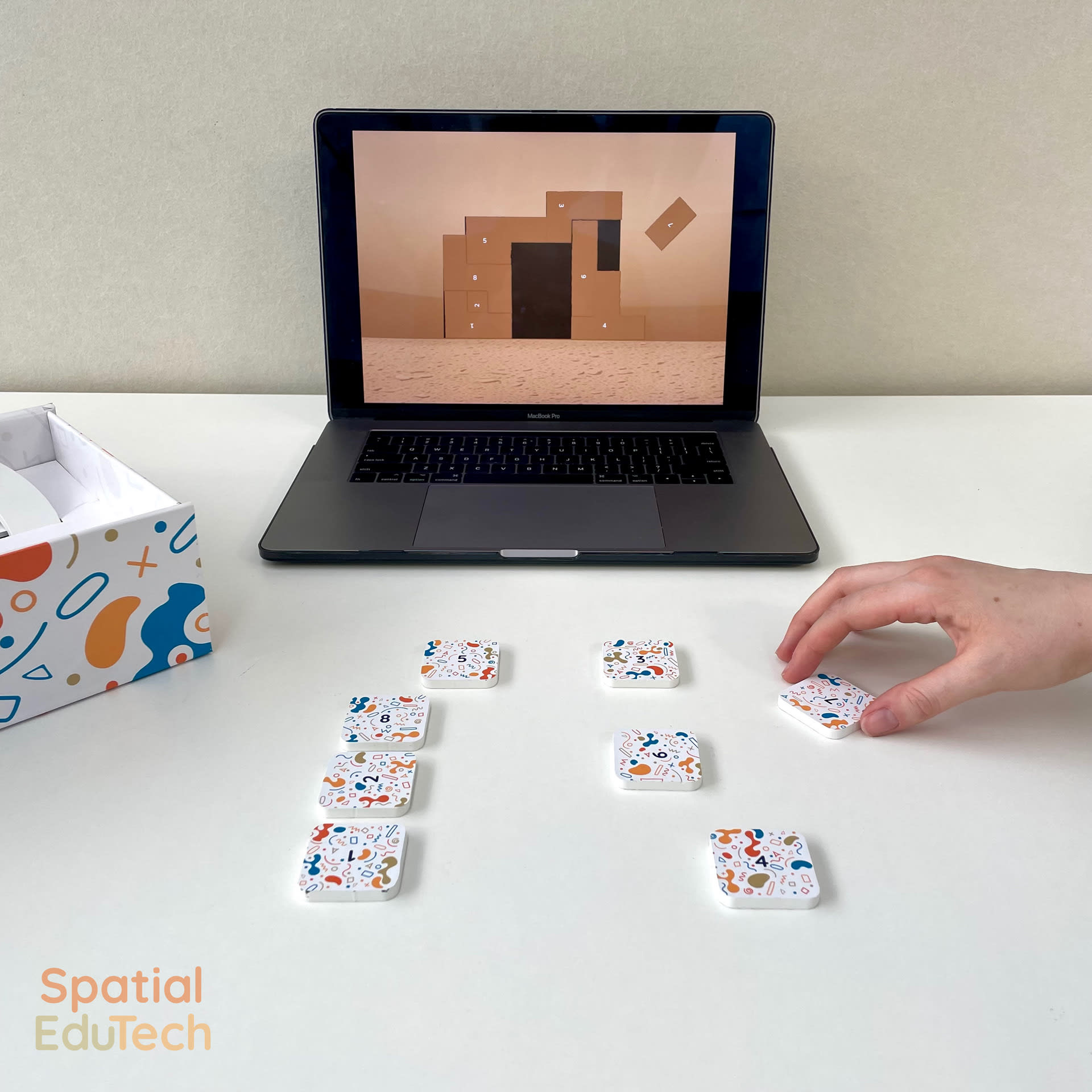 A tiling puzzle on a laptop with physical game pieces in front, with a hand positioning a piece to solve the digital puzzle.